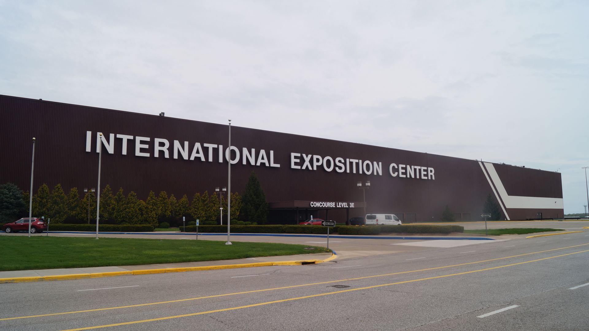 Large brown building labeled "INTERNATIONAL EXPOSITION CENTER" with green bushes and trees in front, a road and parking area, and overcast sky above.