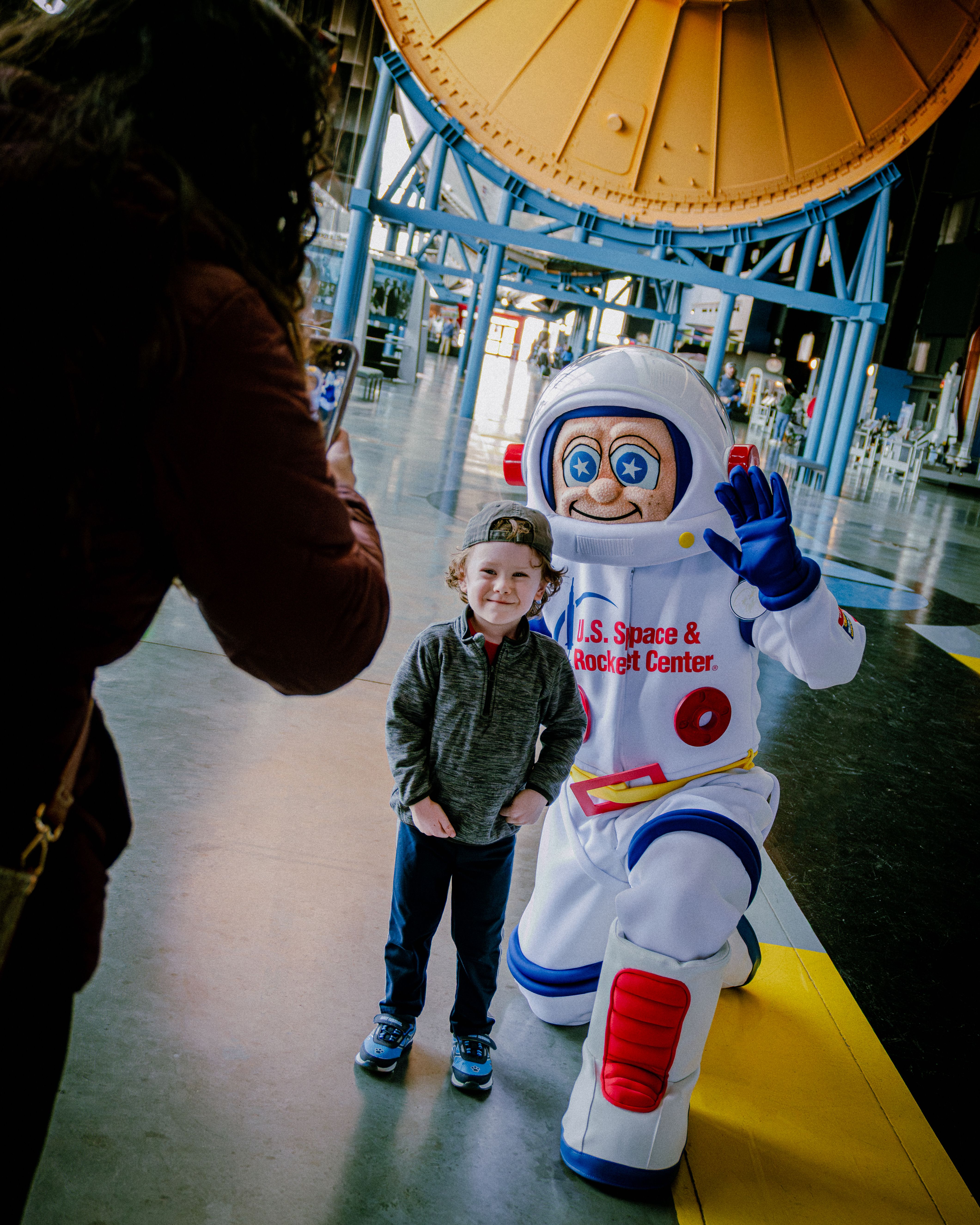 Young boy in a gray jacket poses with a cheerful astronaut mascot in a white suit labeled "U.S. Space & Rocket Center" inside a bright hall with blue beams and a large yellow circular backdrop.