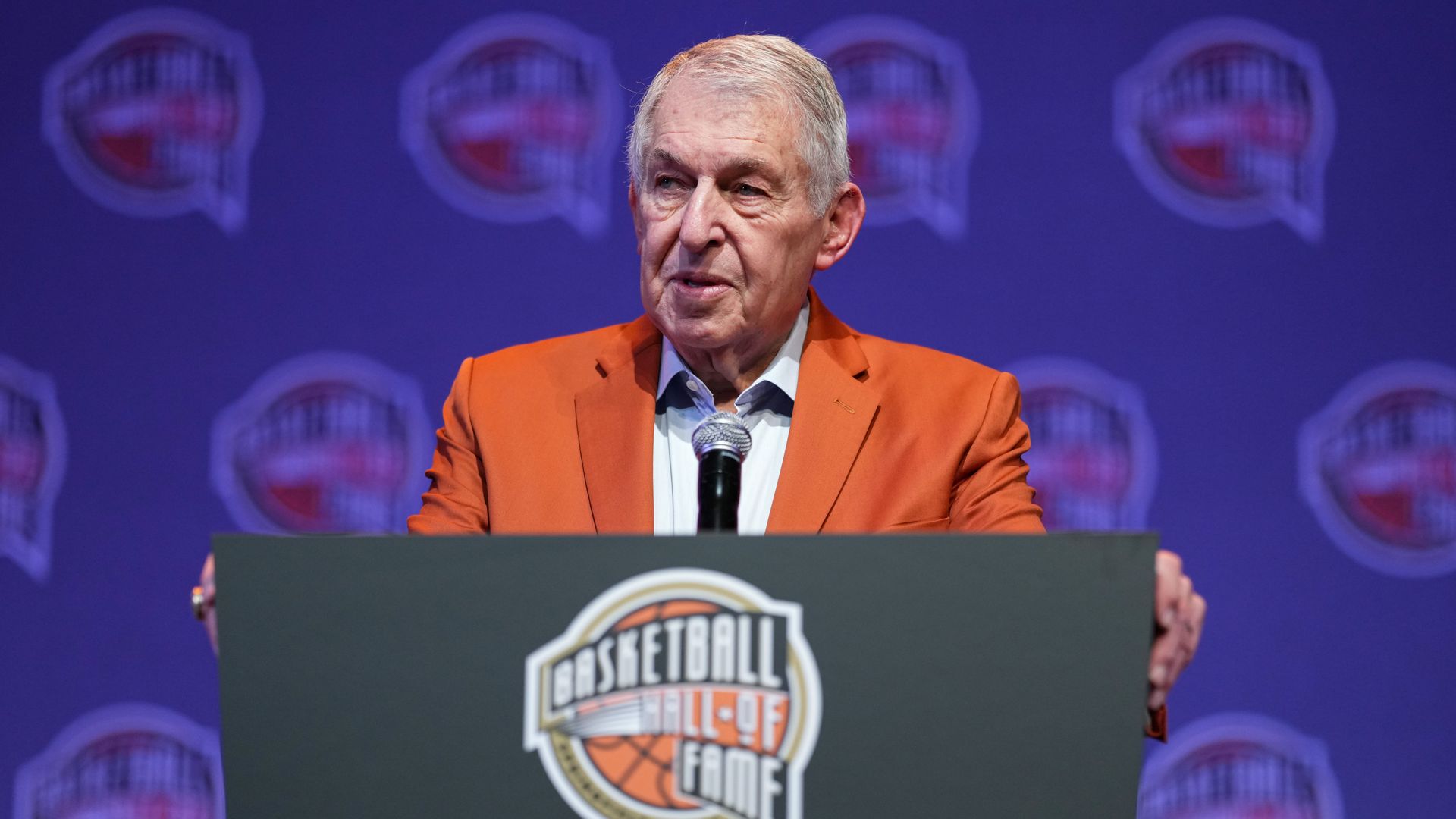 A man in an orange suit jacket and white shirt stands speaks into a microphone on a lectern, with his hands on the sides; a Basketball Hall of Fame logo is on the front of the lectern, and the same logo adorns the background.