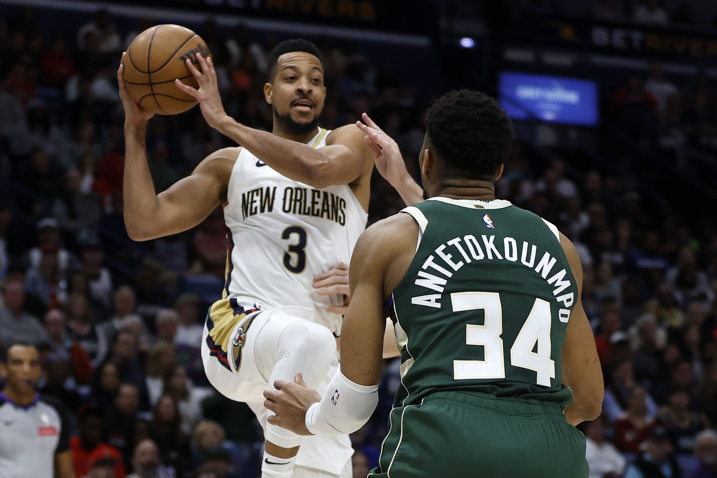 CJ McCollum leaps toward Giannis Antetokounmpo, whose back is to the camera, with a basketball.