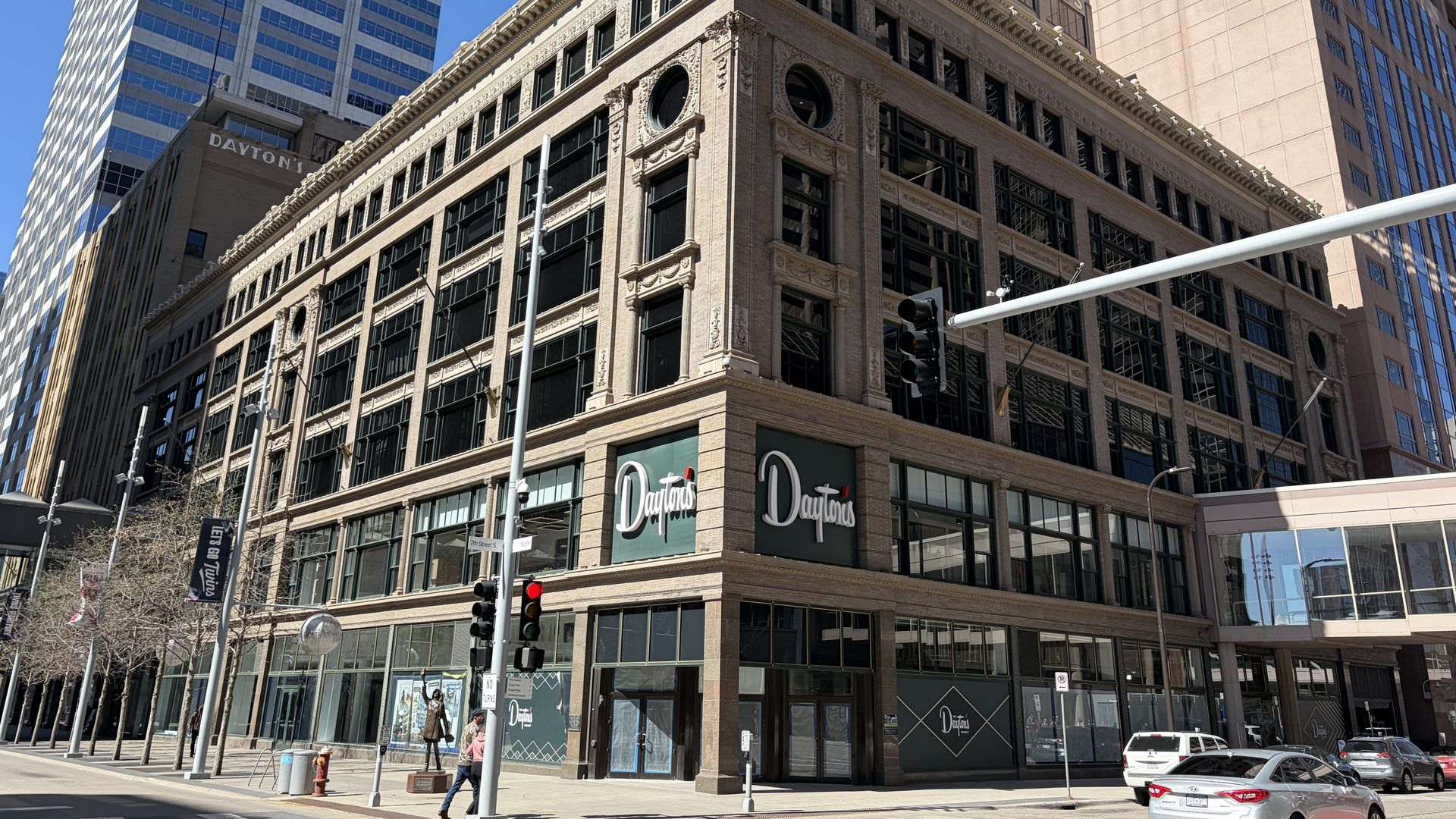 Urban city street scene with a beige brick building in the foreground and modern glass towers behind. Daytons storefronts line the ground floor under blue sky; pedestrians and a car cross the street.