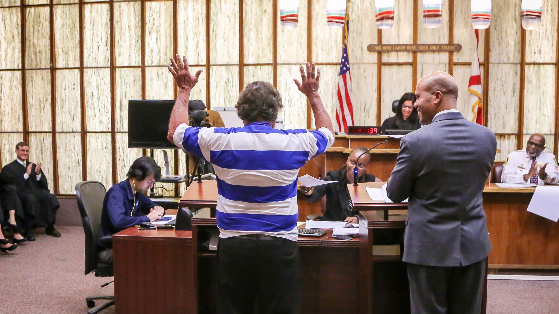 In this image, a person in a courtroom raises their hands