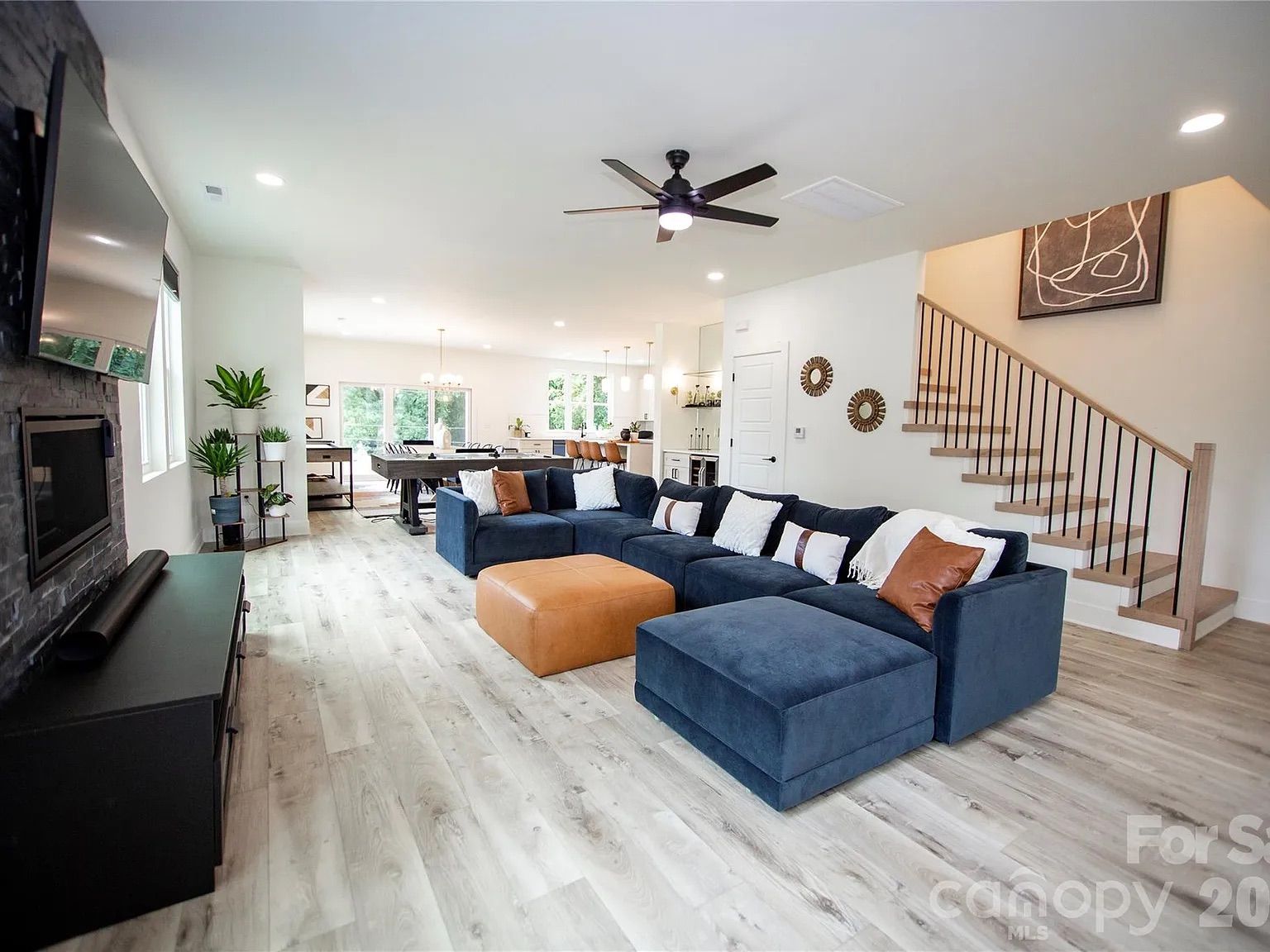 Open living area with a navy L-shaped sofa, white and orange pillows, an orange ottoman, and a blue pouf. Light wood floors, staircase with black rails, fireplace and TV, plants, and large windows.