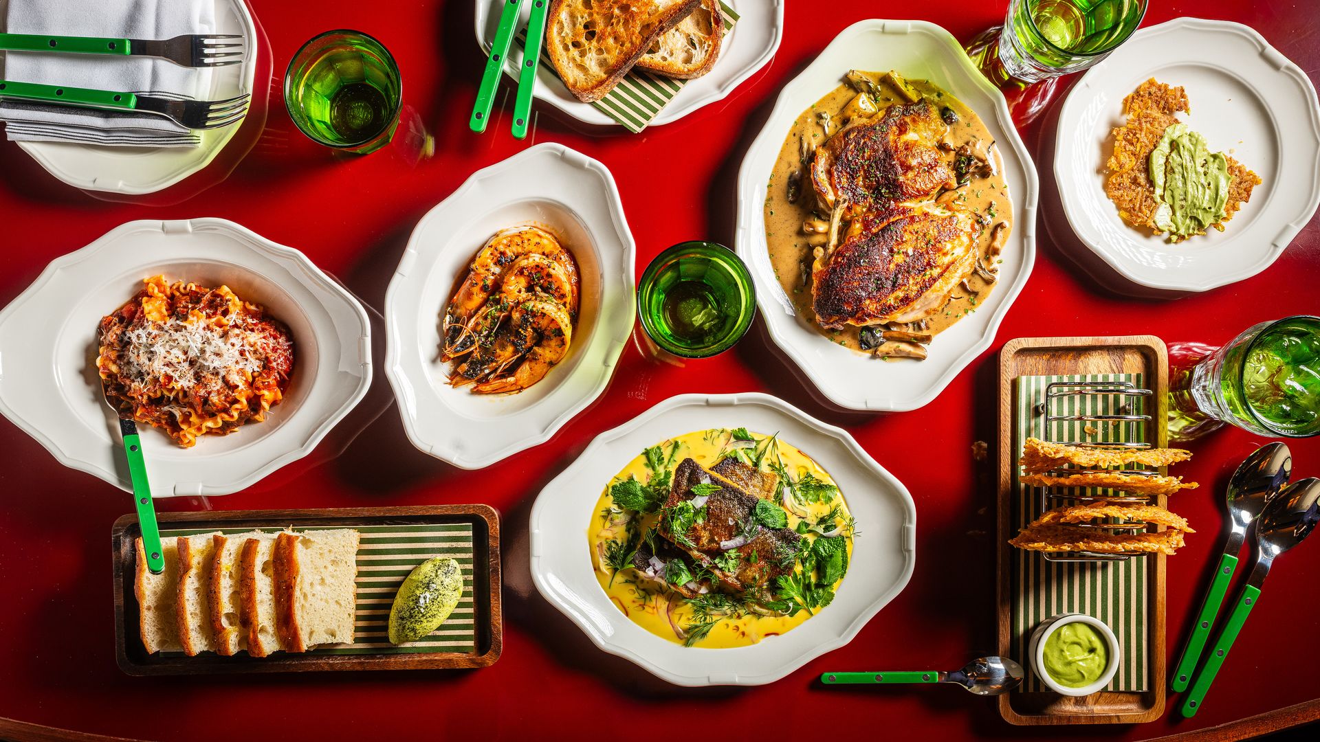 Top-down view of a red table set with white plates, green-handled cutlery, and green glassware. Dishes include roasted chicken in creamy sauce, pasta, avocado-topped toast, and slices of bread.