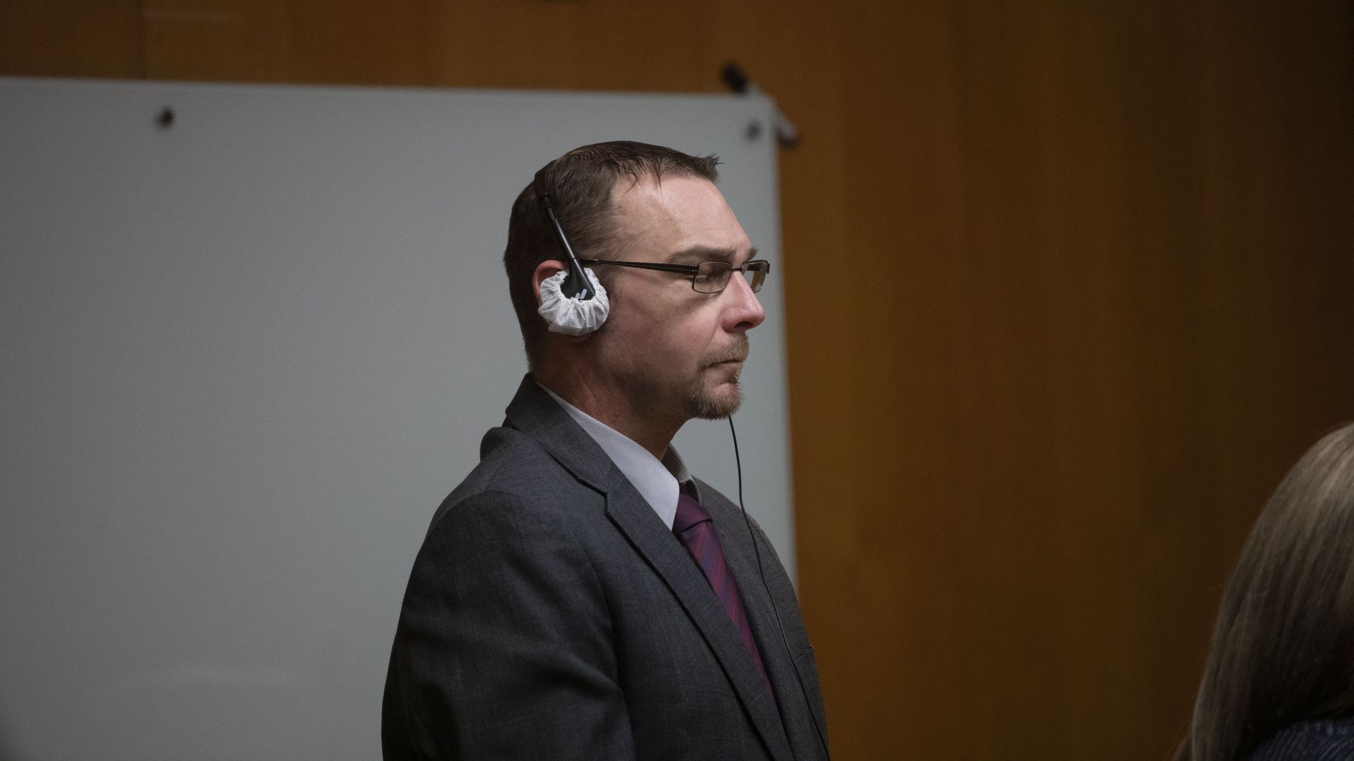 James Crumbley stands wearing a gray suit and burgundy tie with headphones covering his ears. He is wearing glasses and there is a wood wall behind him. 