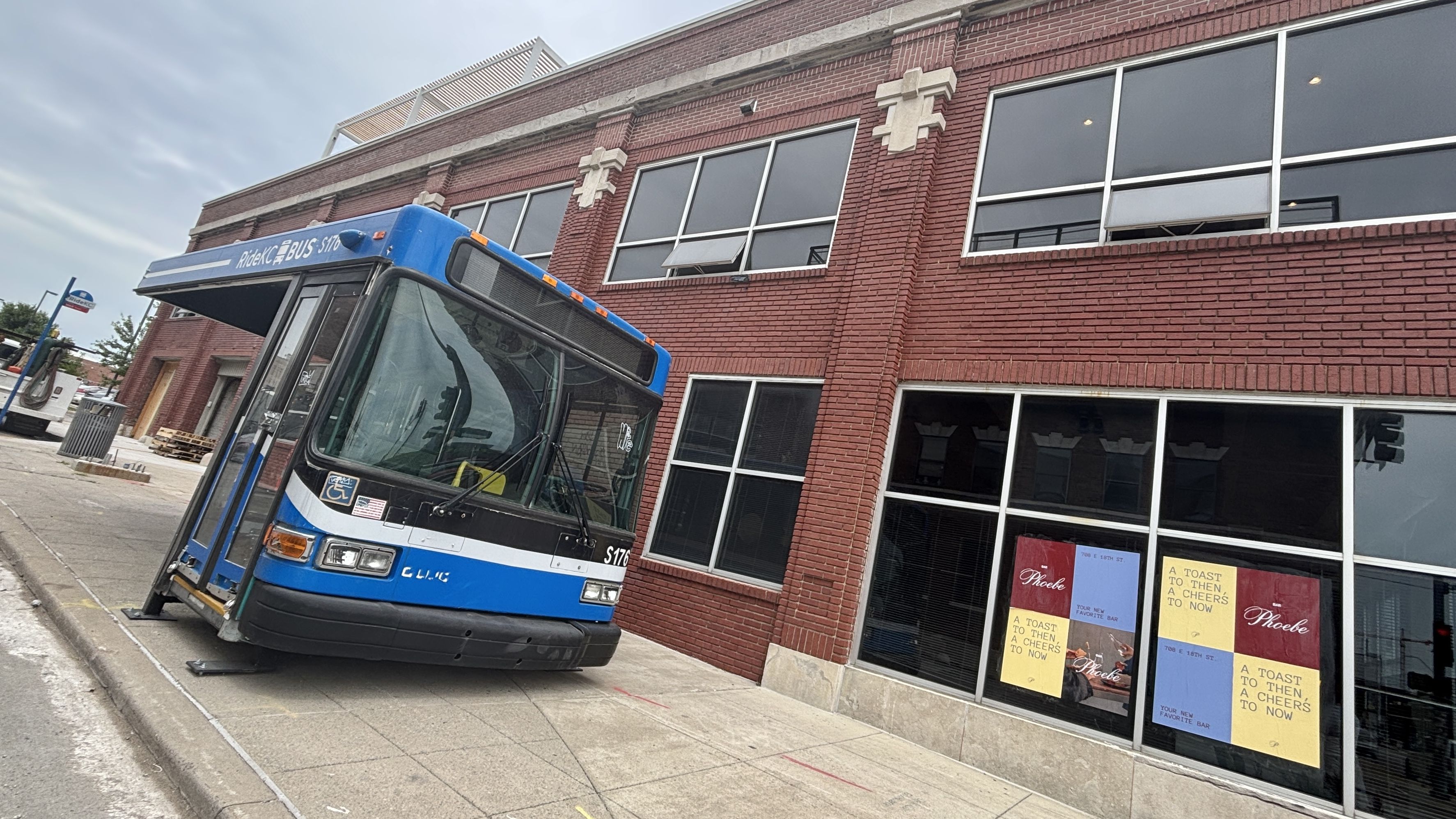 Blue urban transit bus labeled RideKC Bus sits at an angle beside a red brick building with large windows and decorative stone crosses; storefront posters in the windows; cloudy sky.