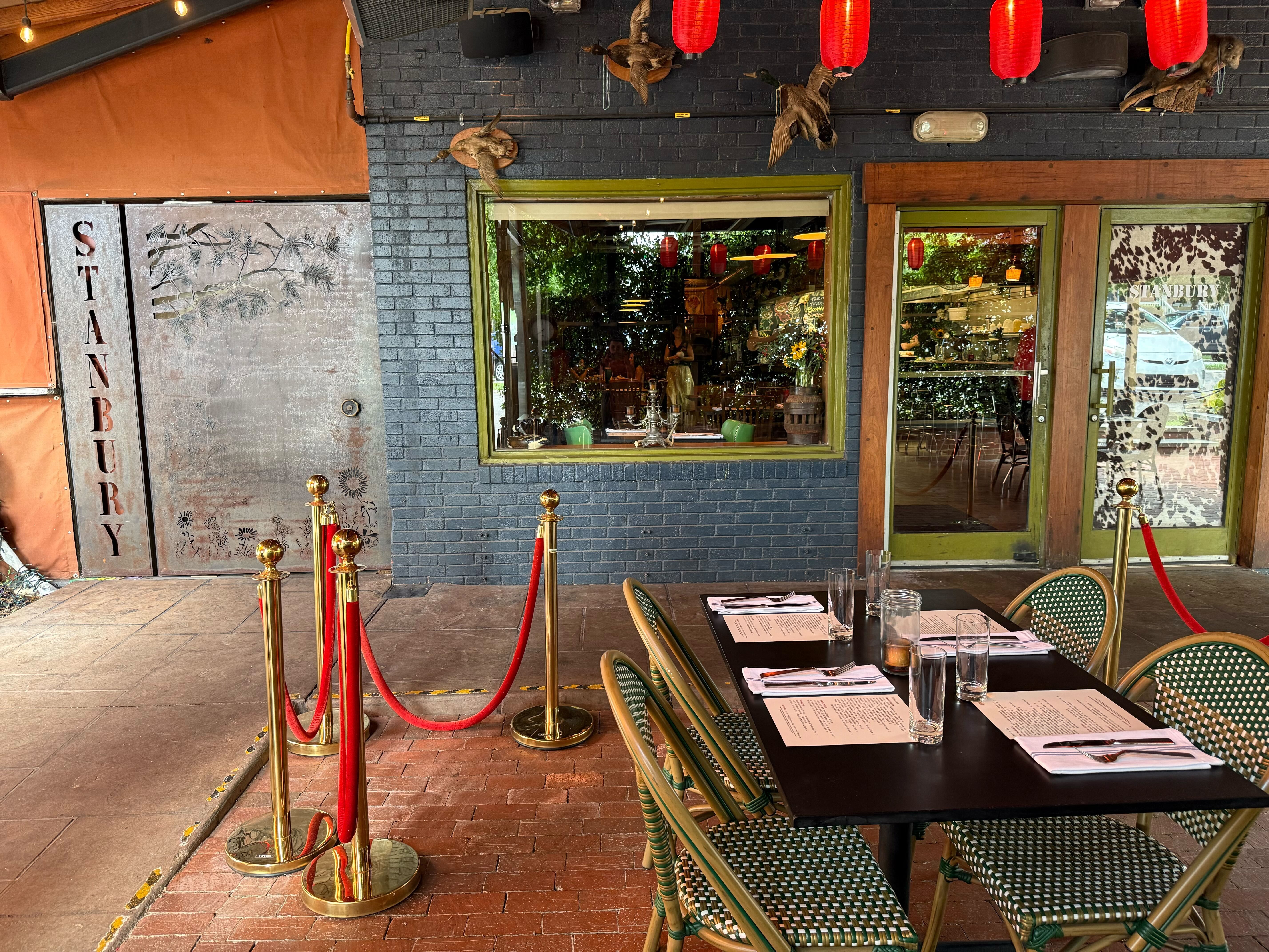 Outdoor dining area of Stanbury restaurant with a table set for four, green woven chairs, red lanterns hanging, black brick wall, and a metal door with Stanbury text.