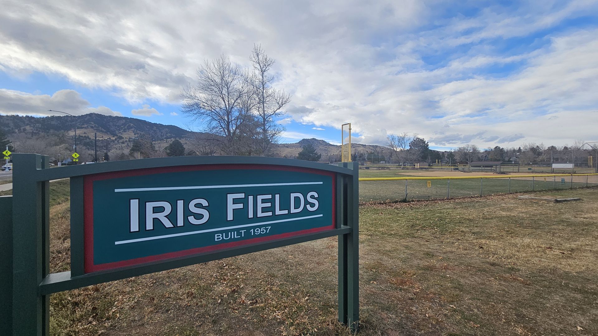 Green and red sign reading "IRIS FIELDS BUILT 1957" in front of a fenced baseball field with yellow fencing and brown grass under a partly cloudy blue sky with hills in the background.