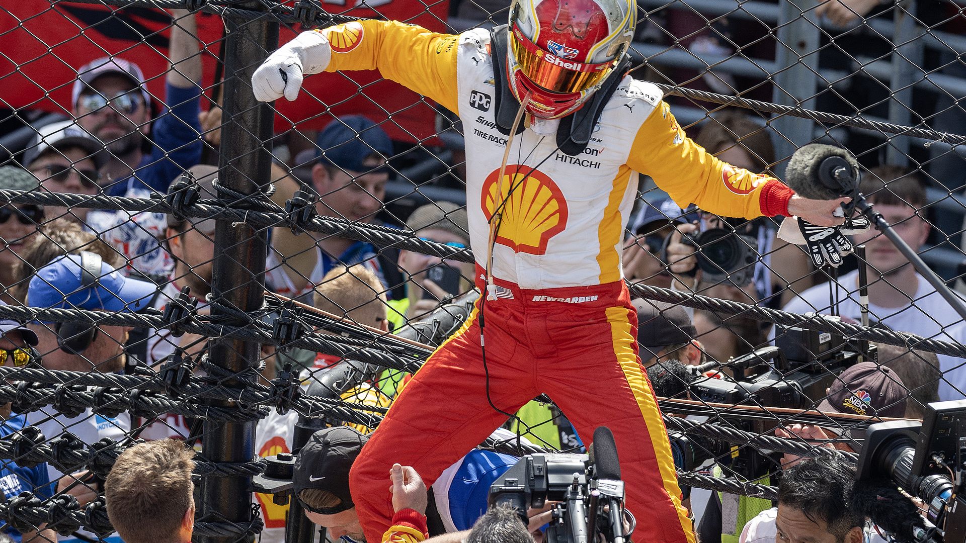 A driver in a fire suit jumping down from the fence in front of cameras