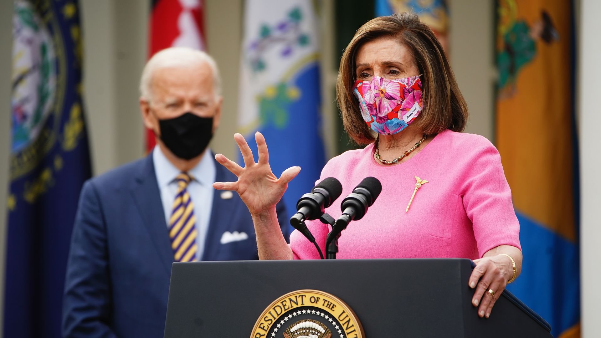Picture of Nancy Pelosi speaking before a podium and President Biden in the background