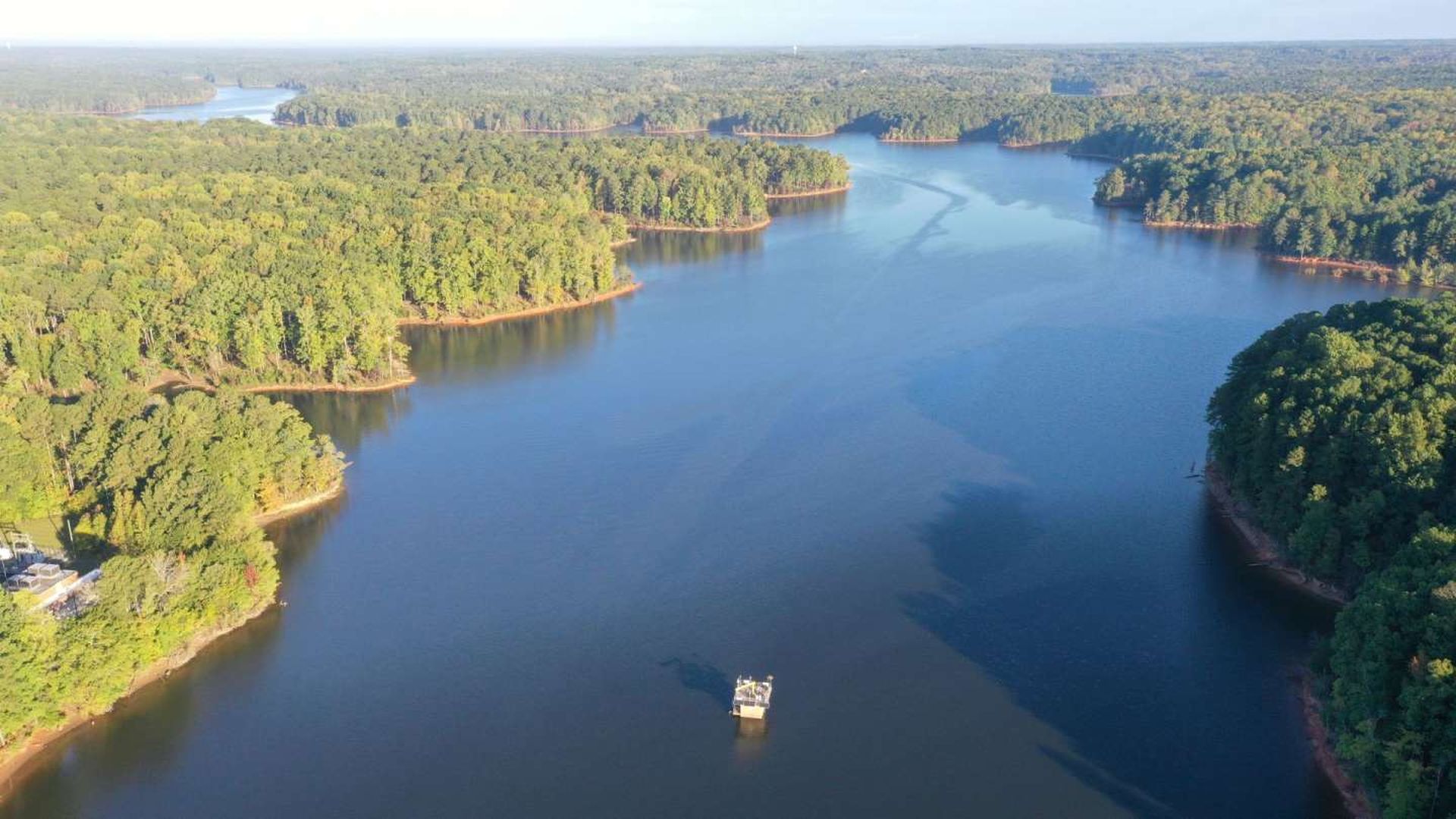 Aerial view of a winding river bordered by dense green forests. Calm blue water snakes between tree-lined shores, with a small boat near the center.