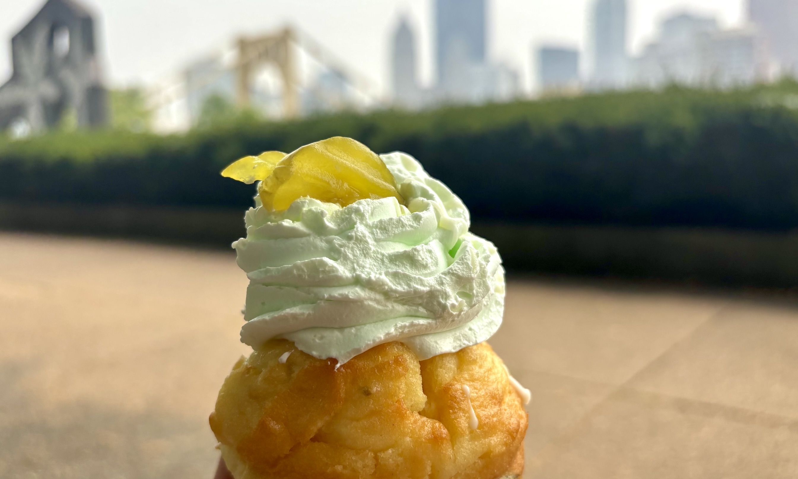 Close-up of a stacked pastry topped with swirled whipped cream and a yellow lemon garnish, set against a blurred city skyline and green hedge background.