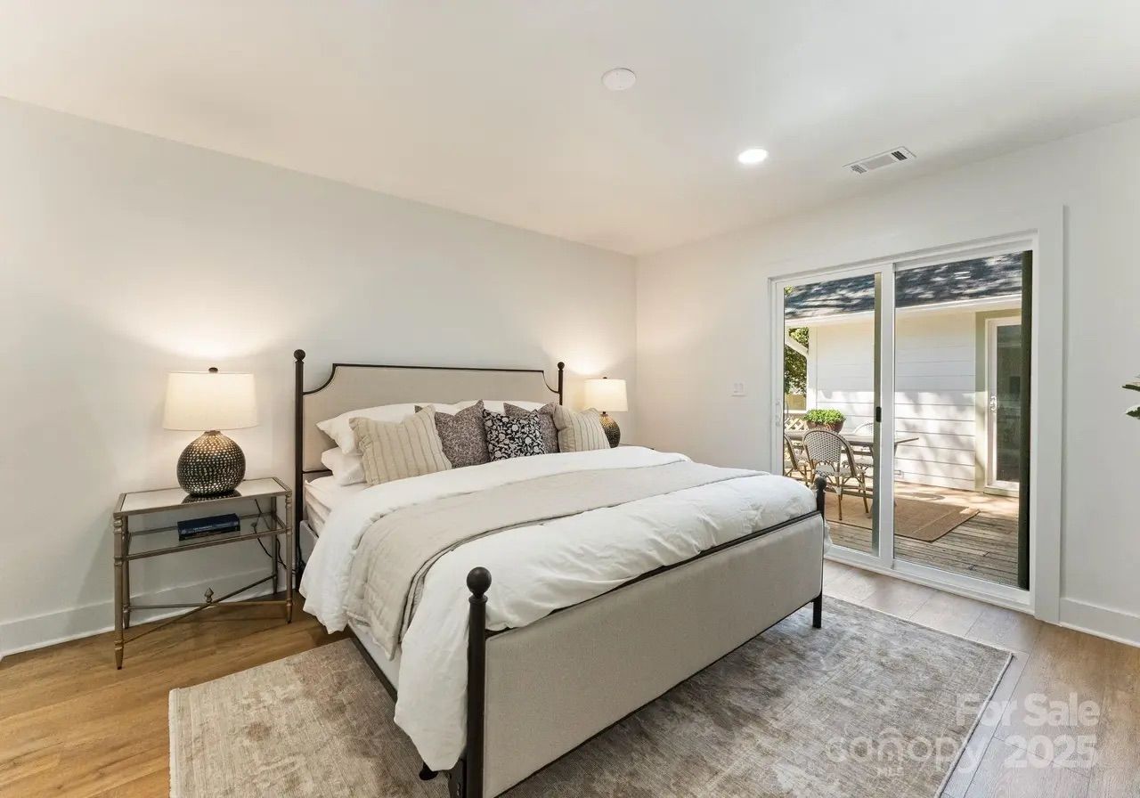 Bright bedroom with a beige upholstered bed, white and patterned pillows, wooden floors, side tables with lamps, and sliding glass doors opening to a wooden patio with chairs.