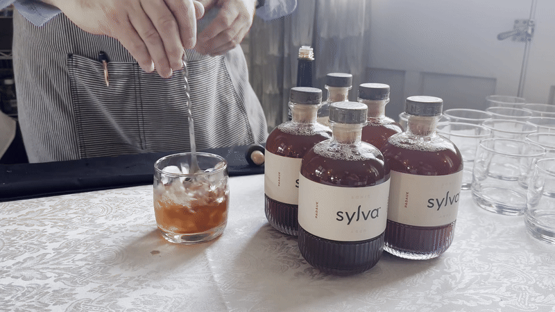 A person in a striped apron stirs a brown drink with ice in a clear glass on a white patterned table. Five bottles labeled "Sylva" filled with the same brown liquid are nearby.