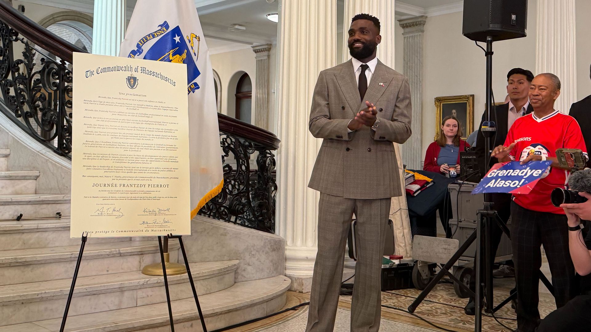 A man in a gray suit speaks at a lectern with a Massachusetts proclamation, beside a white flag and ornate columns; a large speaker and several onlookers are visible.
