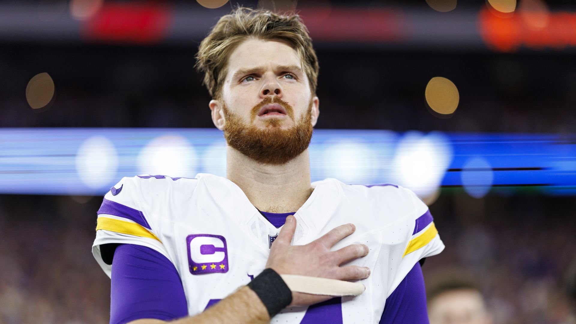 Sam Darnold holds his hand over his heart during the national anthem while wearing a white football jersey and pads