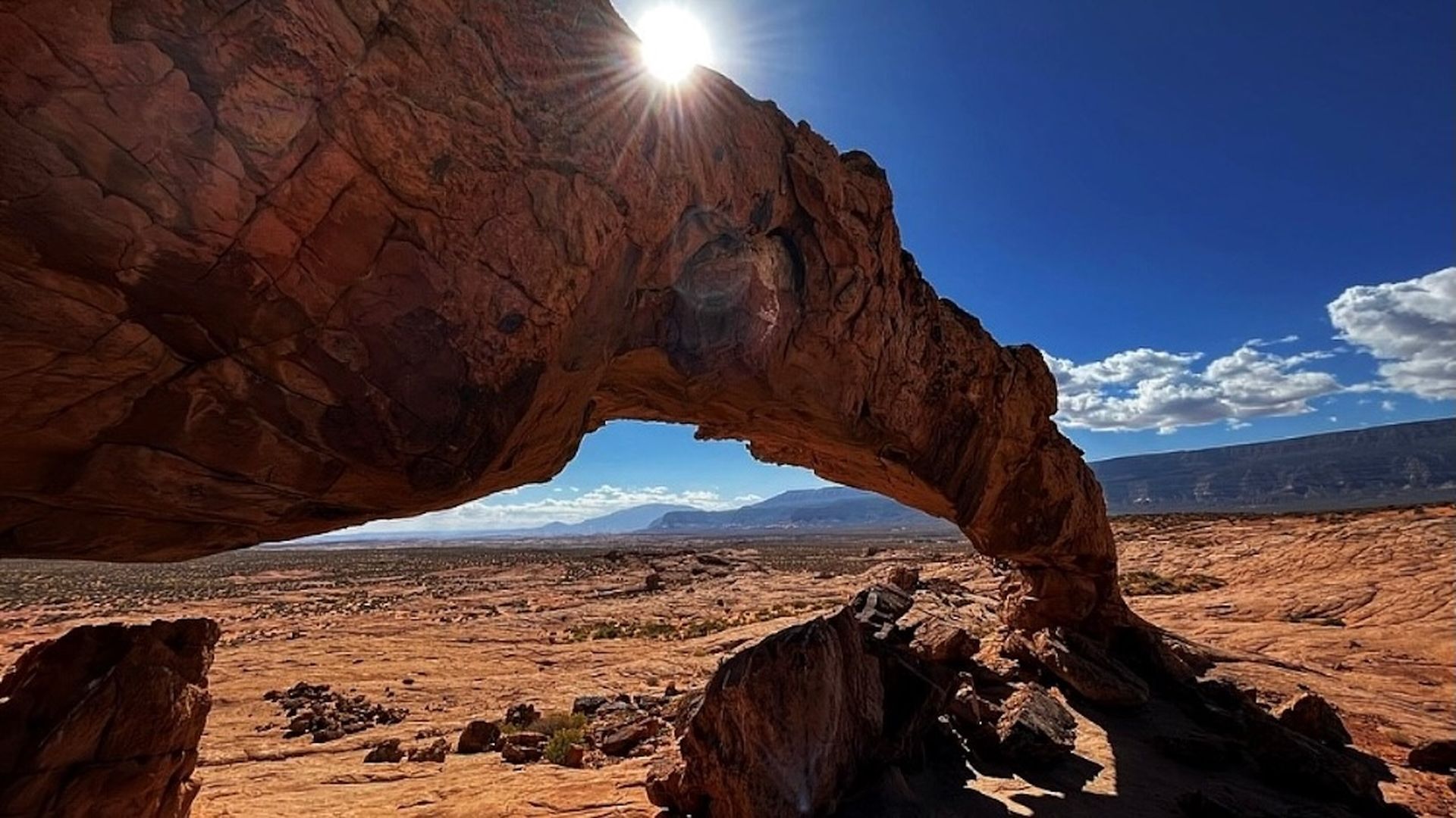 The sun peeks out from behind a stone arch in a redrock desert.