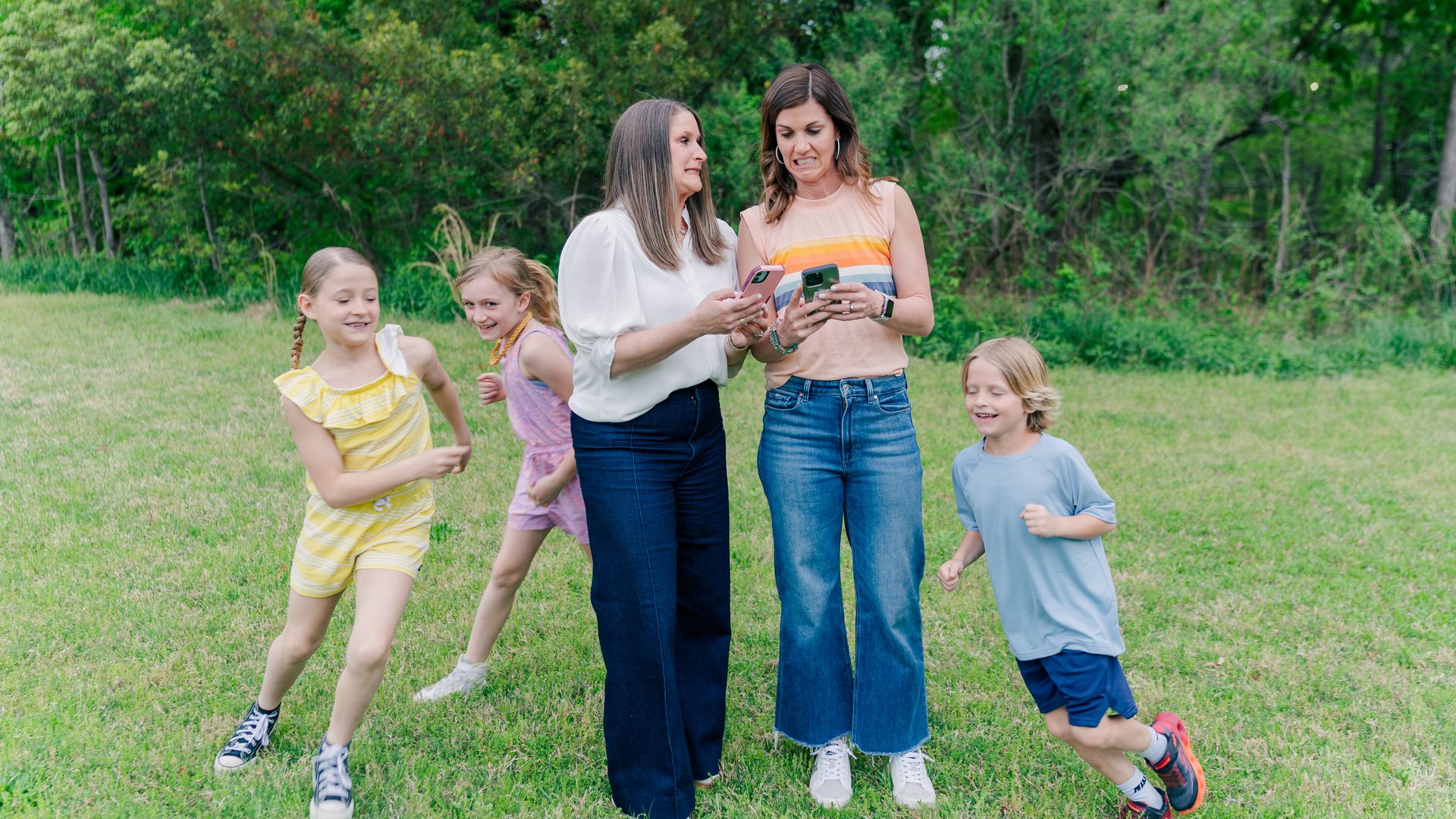 Two women stand in a grassy park with green trees, looking at their phones while several children run and play nearby.