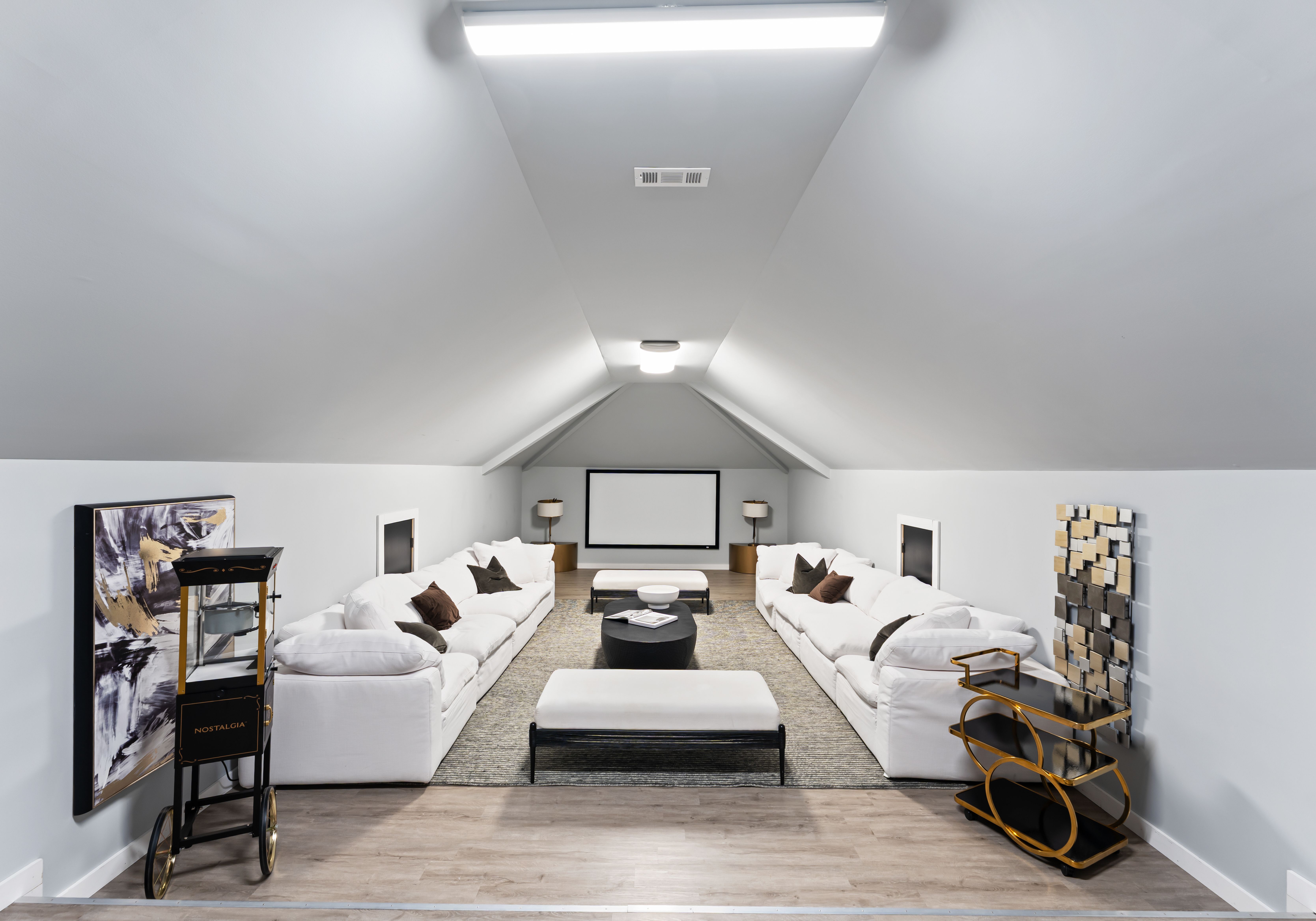 Bright attic living room with slanted gray ceiling, two white sofas facing each other, a black coffee table, white benches, and a projector screen at the end.