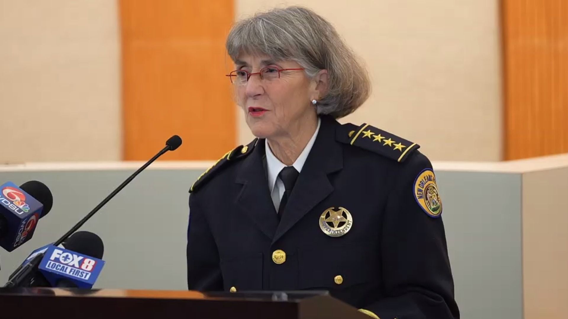 NOPD Superintendent Anne Kirkpatrick speaking at a podium wearing a black police uniform with gold stars and a badge labeled "Superintendent.
