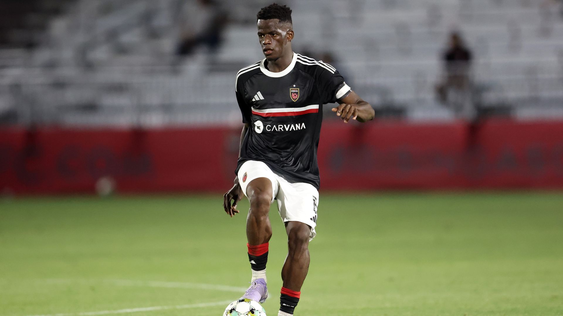 Soccer player in black jersey with white shorts and purple cleats controlling a white and green ball on a grass field, with a blurred stadium background.