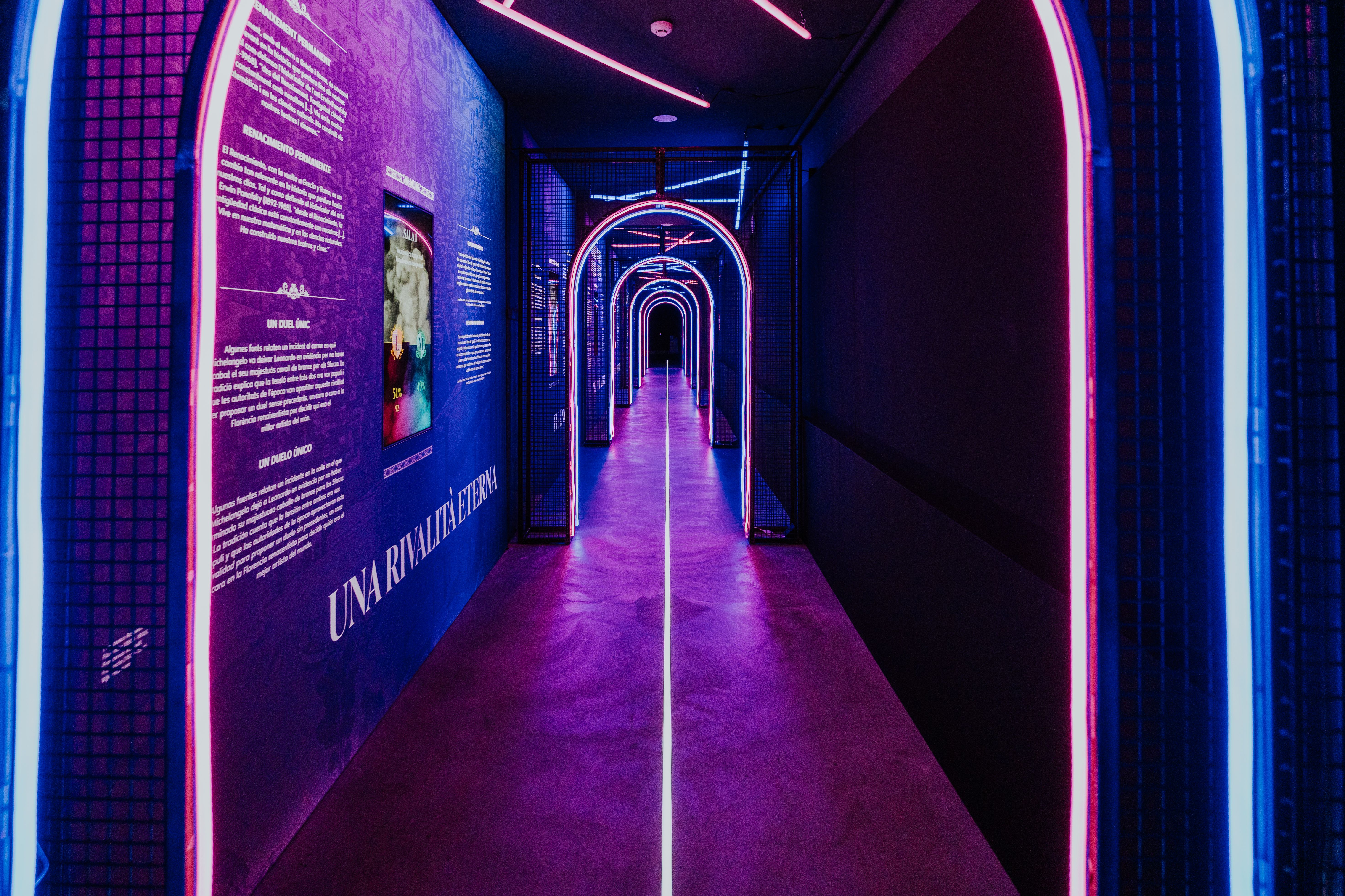 A neon-lit corridor in a museum exhibit: blue left wall with white text panels, a row of glowing pink and blue circular arches forming a tunnel, purple-blue lighting along the floor.