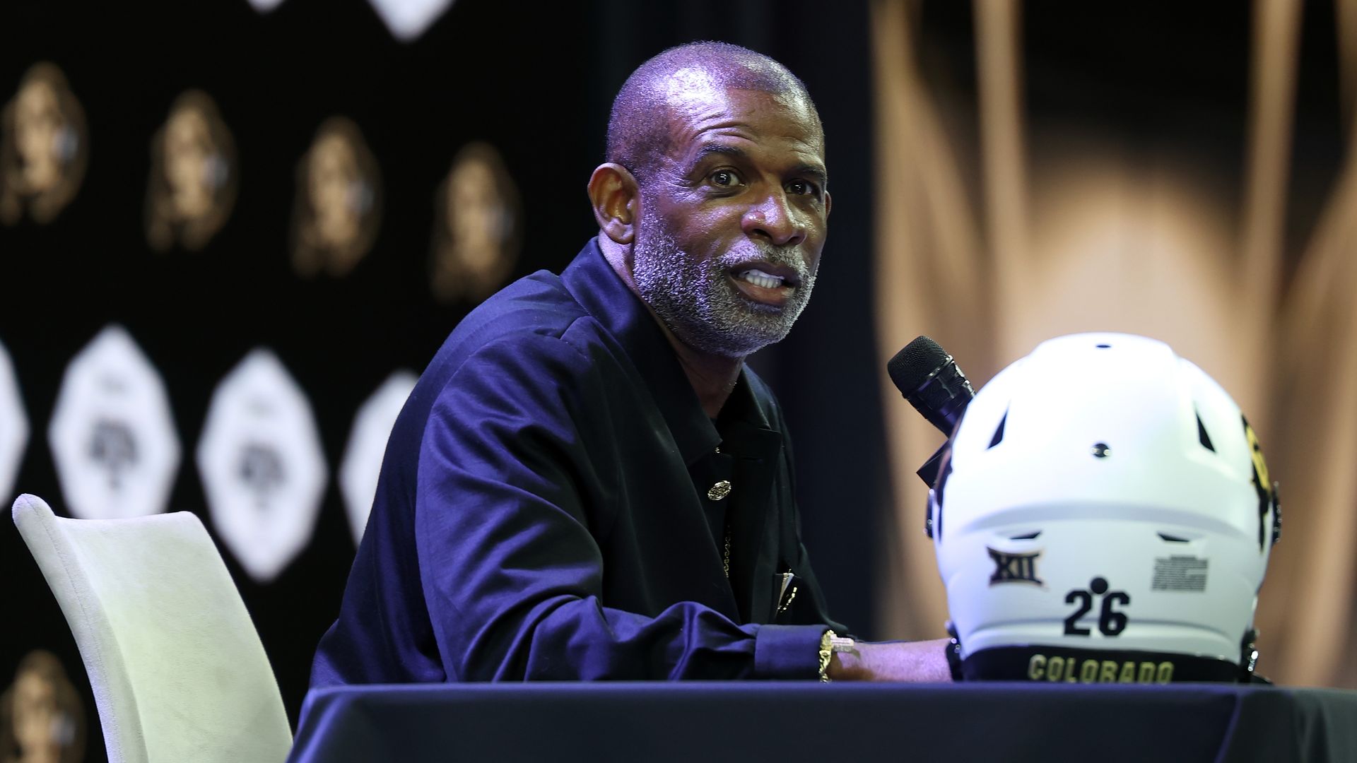 Deion Sanders sits at a table, speaking into a microphone. He wears a dark blue shirt; a white Colorado football helmet with the number 26 rests on the table, against a dark backdrop.