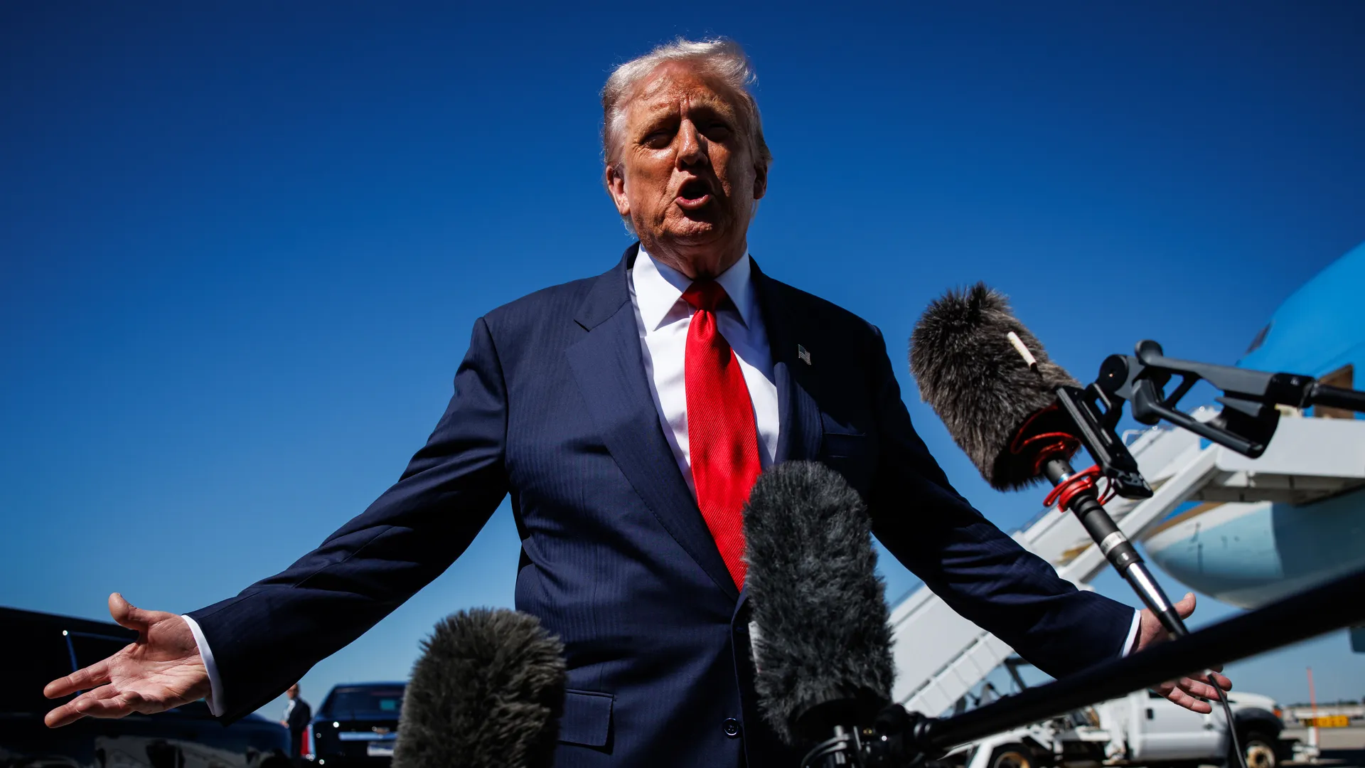 President Trump in dark suit with red tie speaking to multiple microphones outdoors near an airplane with blue sky background.