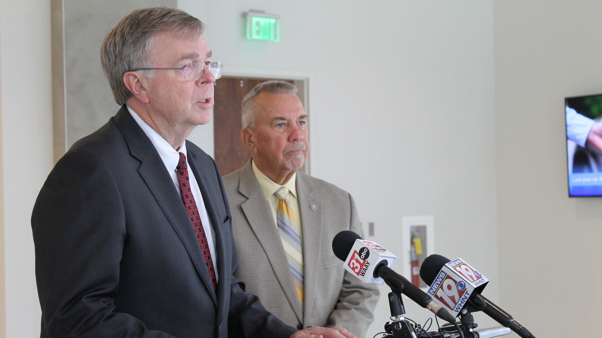 Two older men in suits speak at a press conference with microphones from news channels 31, 19, and 18 in front of them. One wears a dark suit; the other wears a light suit.