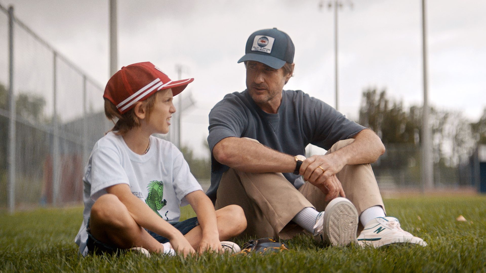 A man in a baseball cap sits next to a young boy on a field