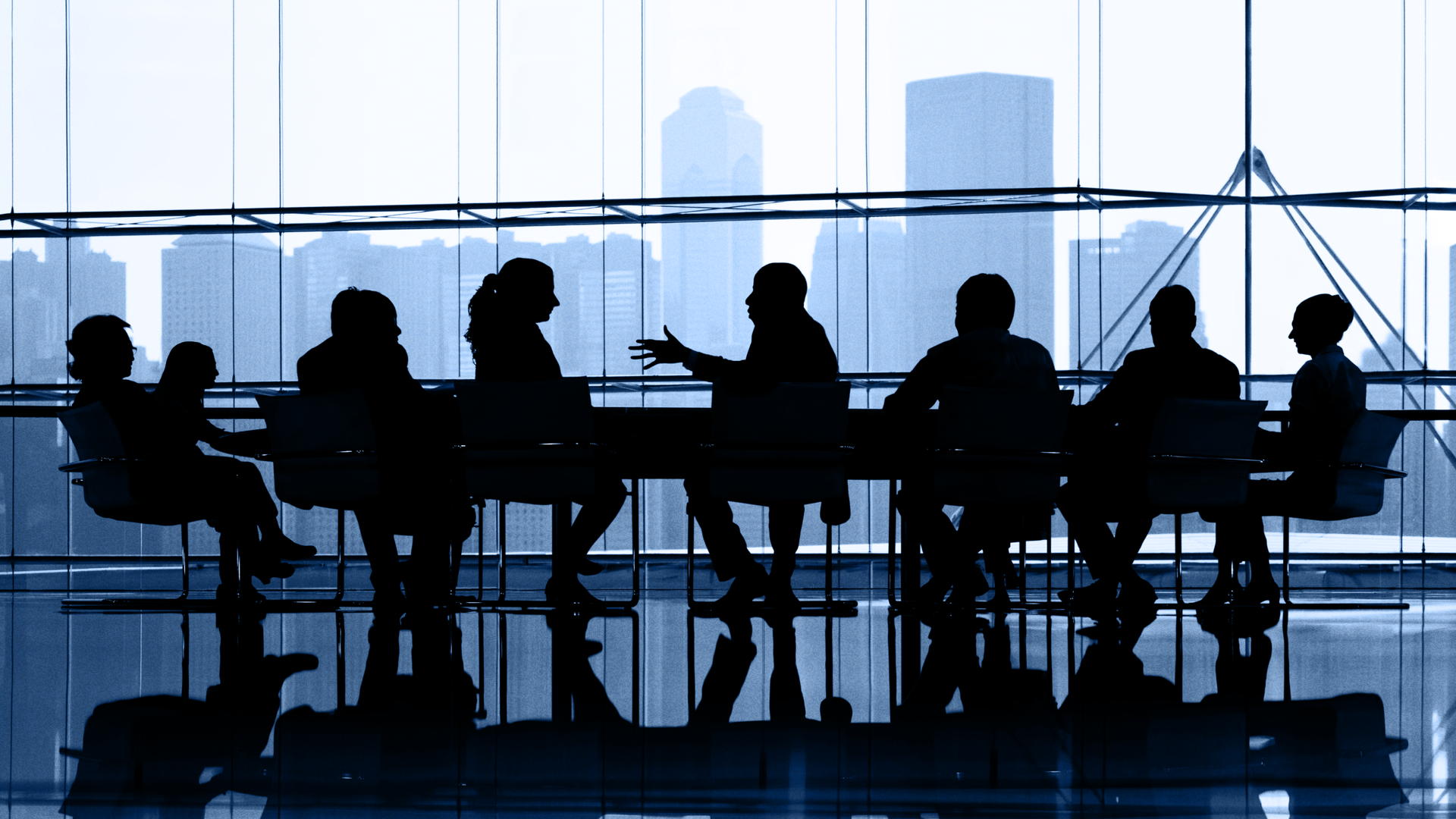 Eight people shrouded in black sit around a conference table, with a skyline image in the backround.
