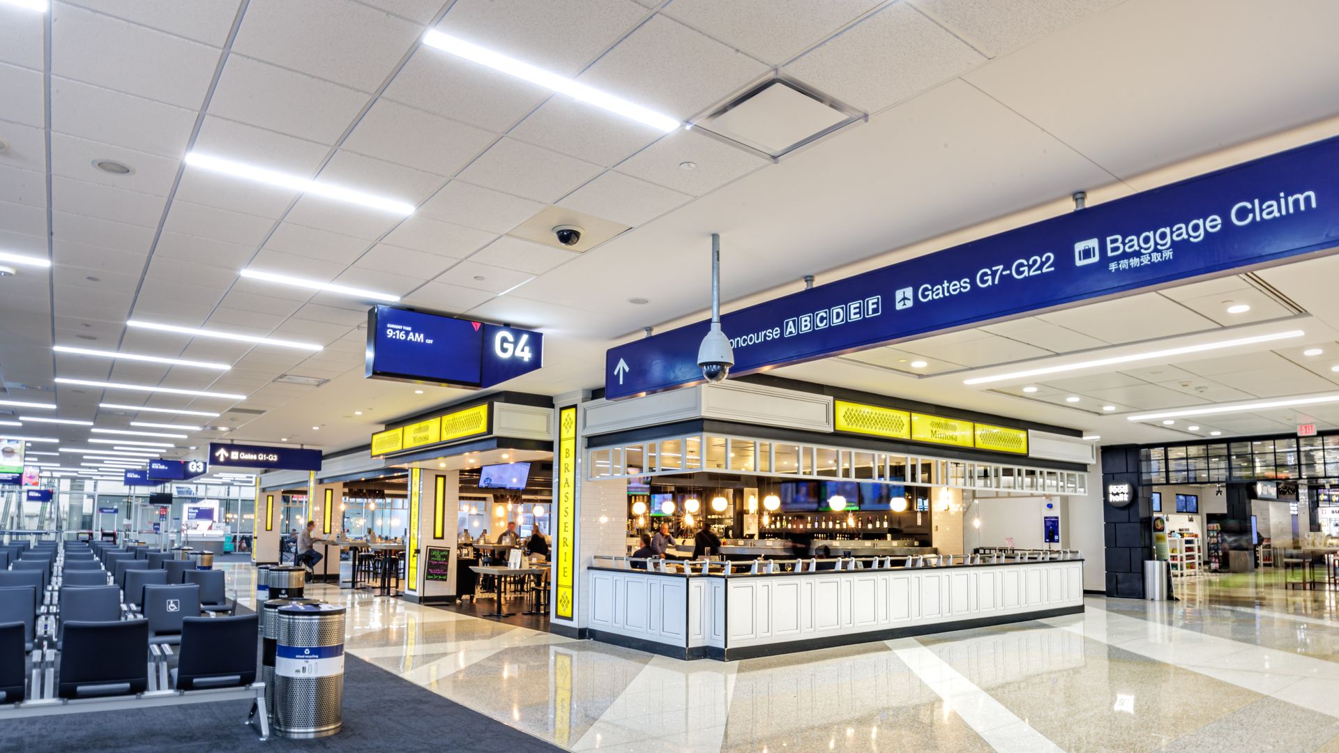 Bright airport concourse with rows of blue seats to the left and a white-bar cafe at center. Overhead blue signs show "G4", "Gates G7-G22", and "Baggage Claim".