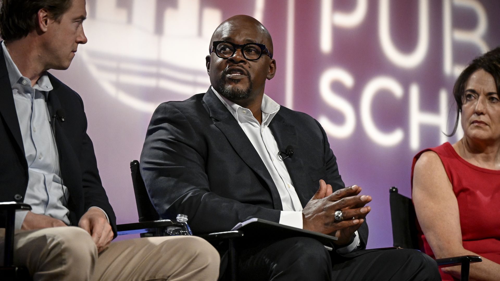 A man in a dark suit and white shirt speaks while seated next to a man and a woman. 