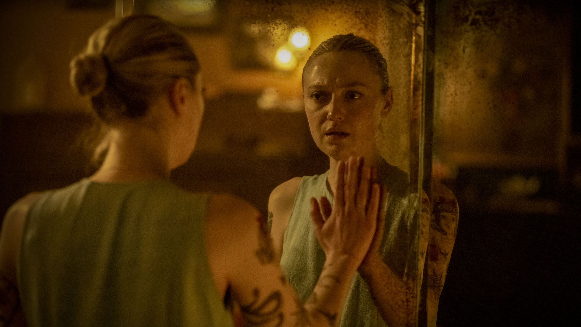Woman with tattoos and light sleeveless top touching a foggy mirror with a concerned expression, warm dim lighting in the room.