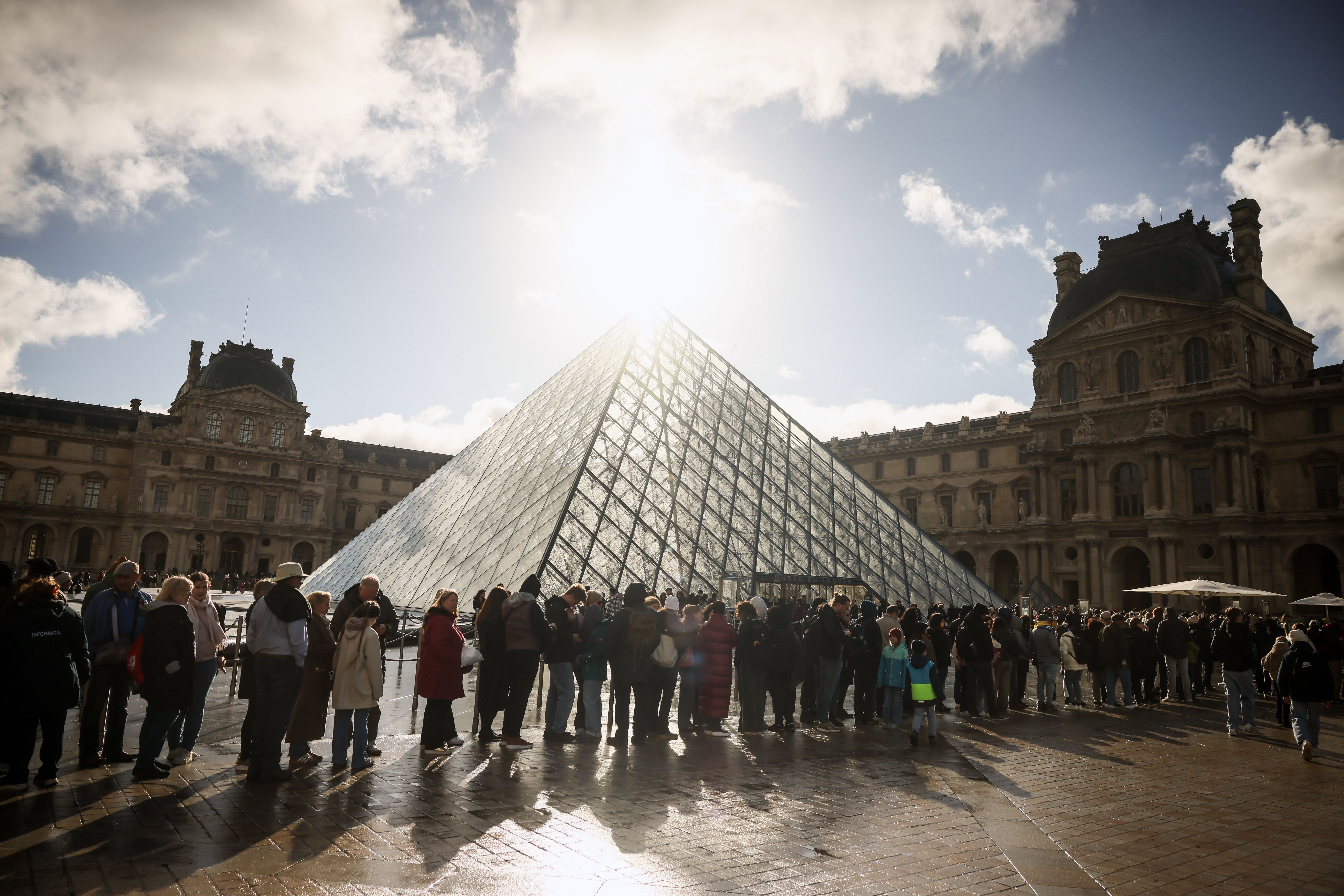 Line of people outside Louvre glass pyramid as sun shines