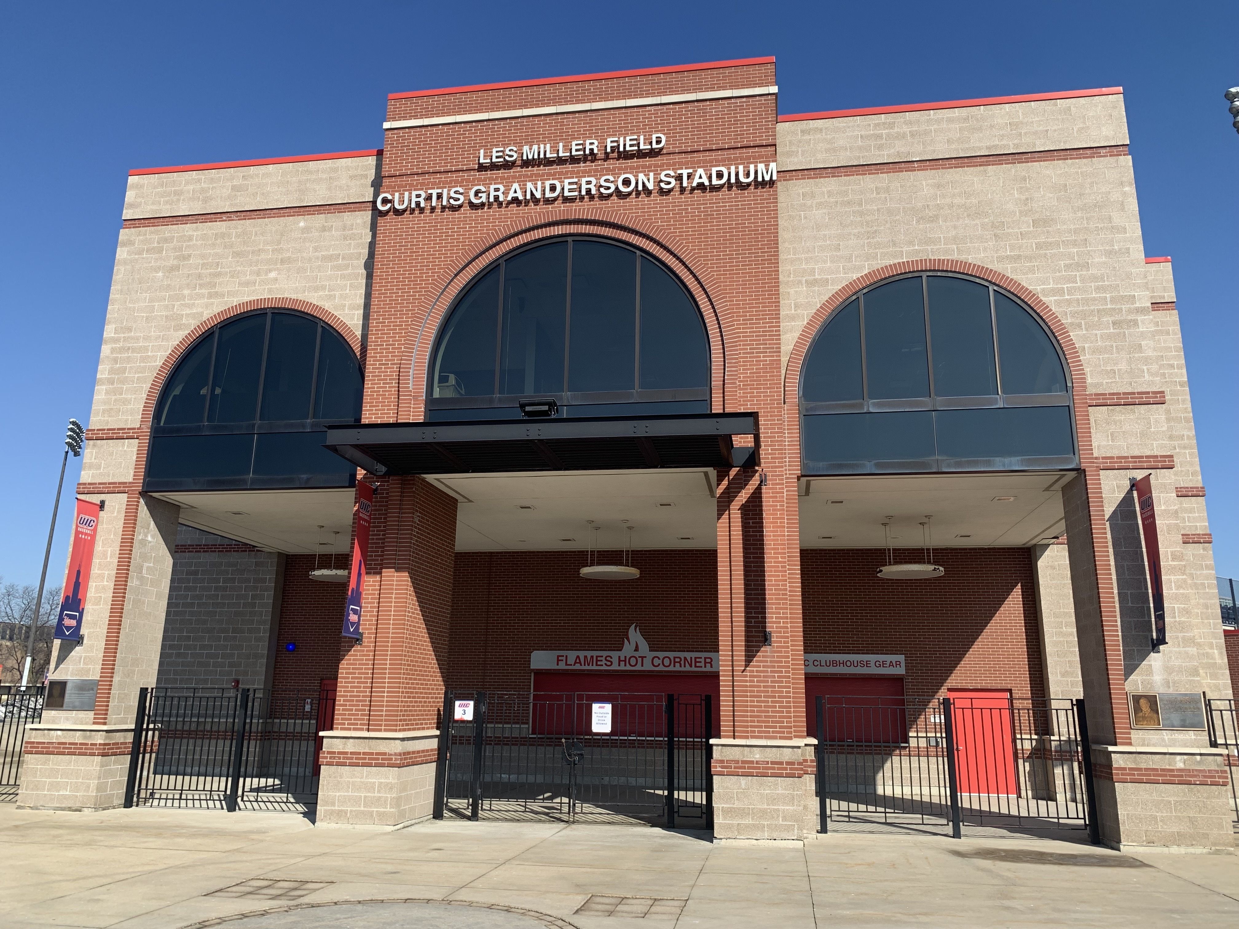 Brick and beige stone entrance to Les Miller Field, Curtis Granderson Stadium with arched windows and red UIC banners against a clear blue sky.