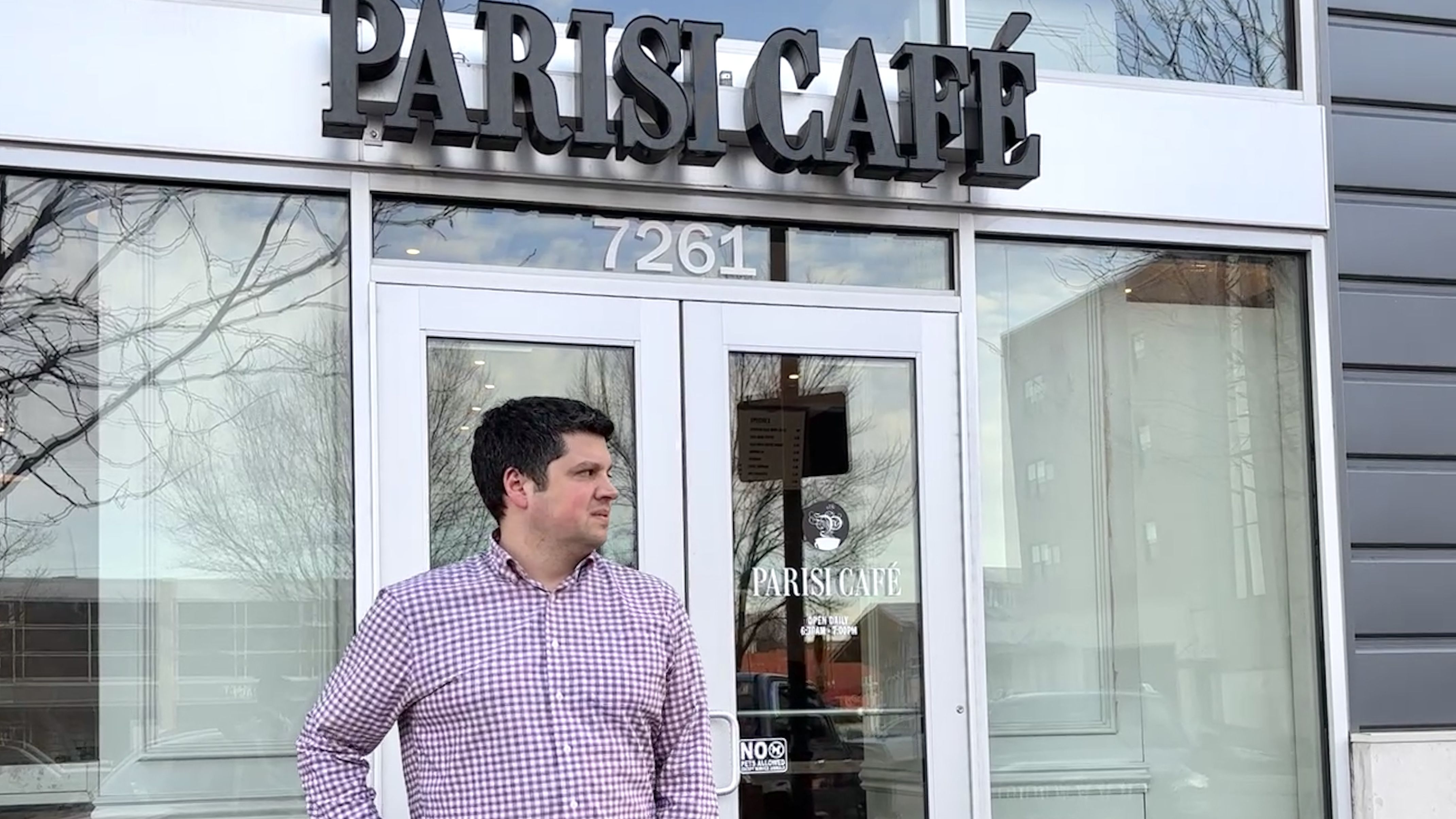 Joe Paris dressed in a purple checkered shirt stands outside Parisi Café with glass doors, building number 7261, and reflections of trees and nearby buildings in the windows.