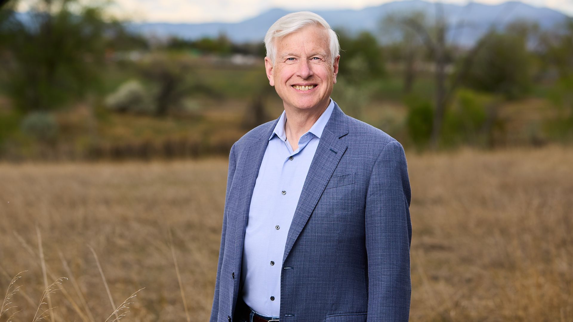Portrait of a smiling older man with white hair, wearing a blue blazer and light blue shirt, standing in a dry field with blurred mountains in the background.