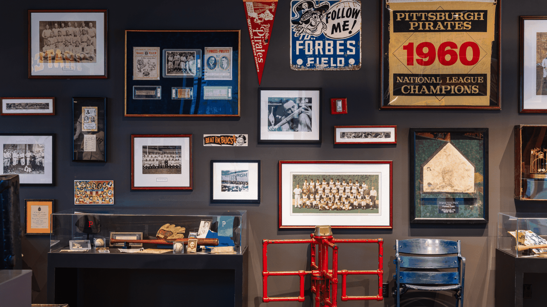 Wall of sports memorabilia with framed photos and posters, a 1960 Pittsburgh Pirates display, vintage signs, and a glass case of baseball gear; a red frame and blue chair in front.