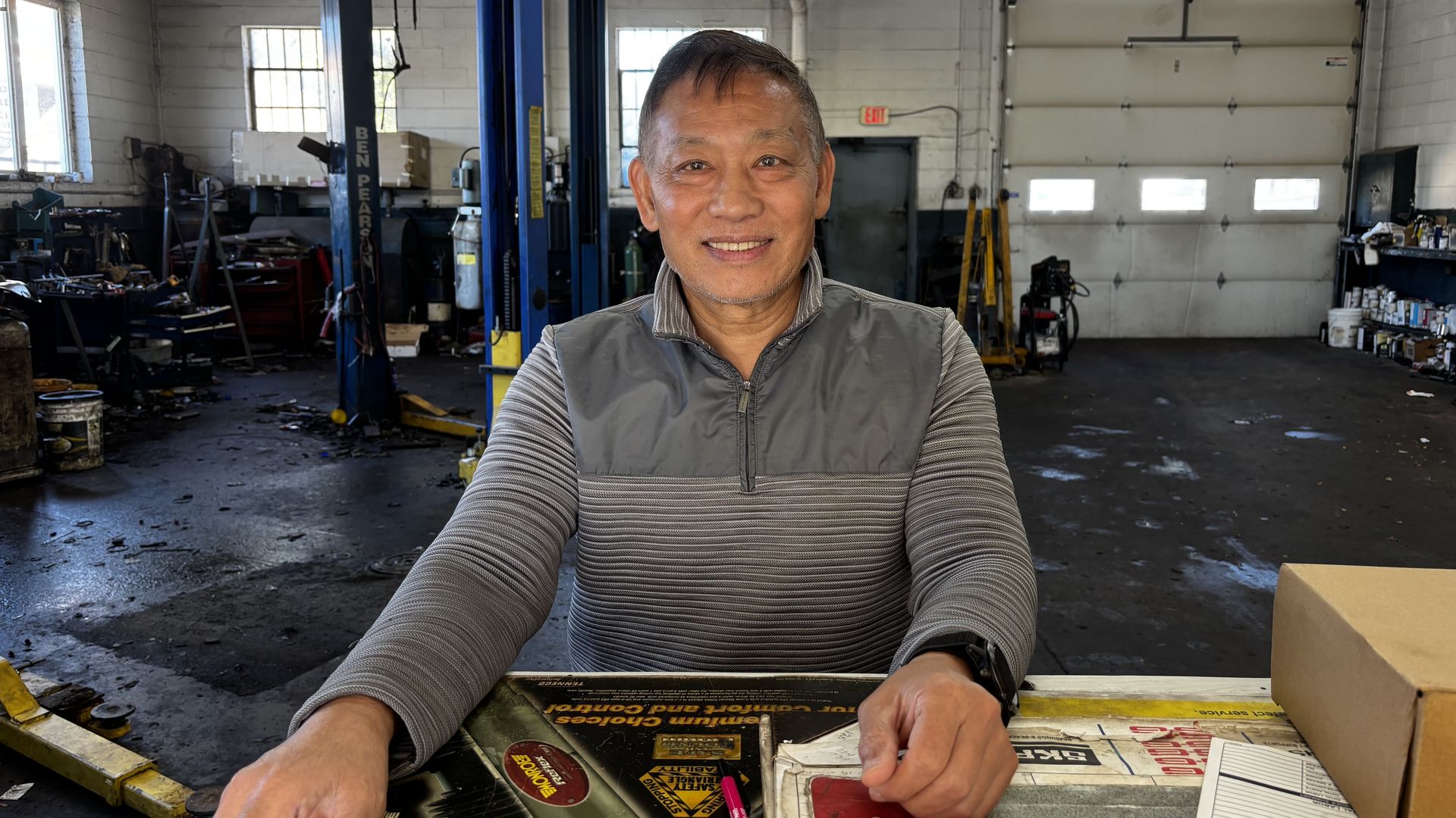 Smiling man in a gray vest over a striped shirt leans over a cluttered auto repair bench inside a busy shop, with a blue car lift, white walls, and a large garage door in the background.