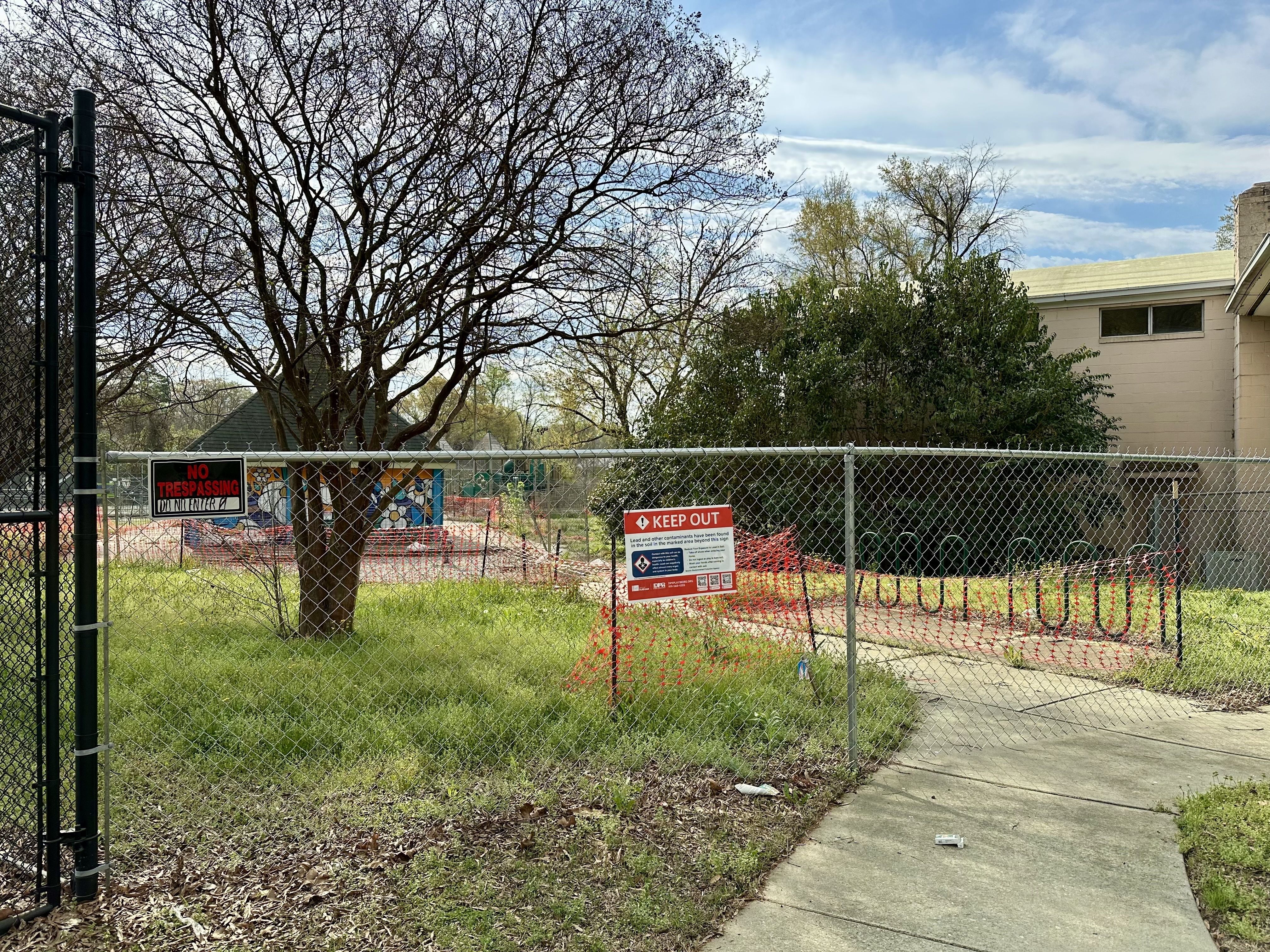 Chain-link fence surrounds a vacant lot with a large bare tree, orange safety netting, and a beige building. Red "KEEP OUT" and "NO TRESPASSING" signs stand near a curved sidewalk.