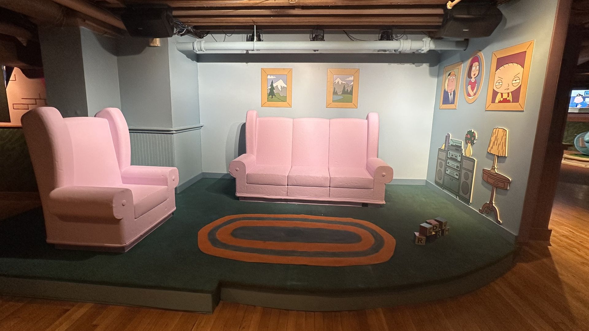 Room setup mimicking "Family Guy" living room with pink couch and chair, green carpet with orange oval, painted wall portraits of characters, and cartoon-style stereo and lamp.