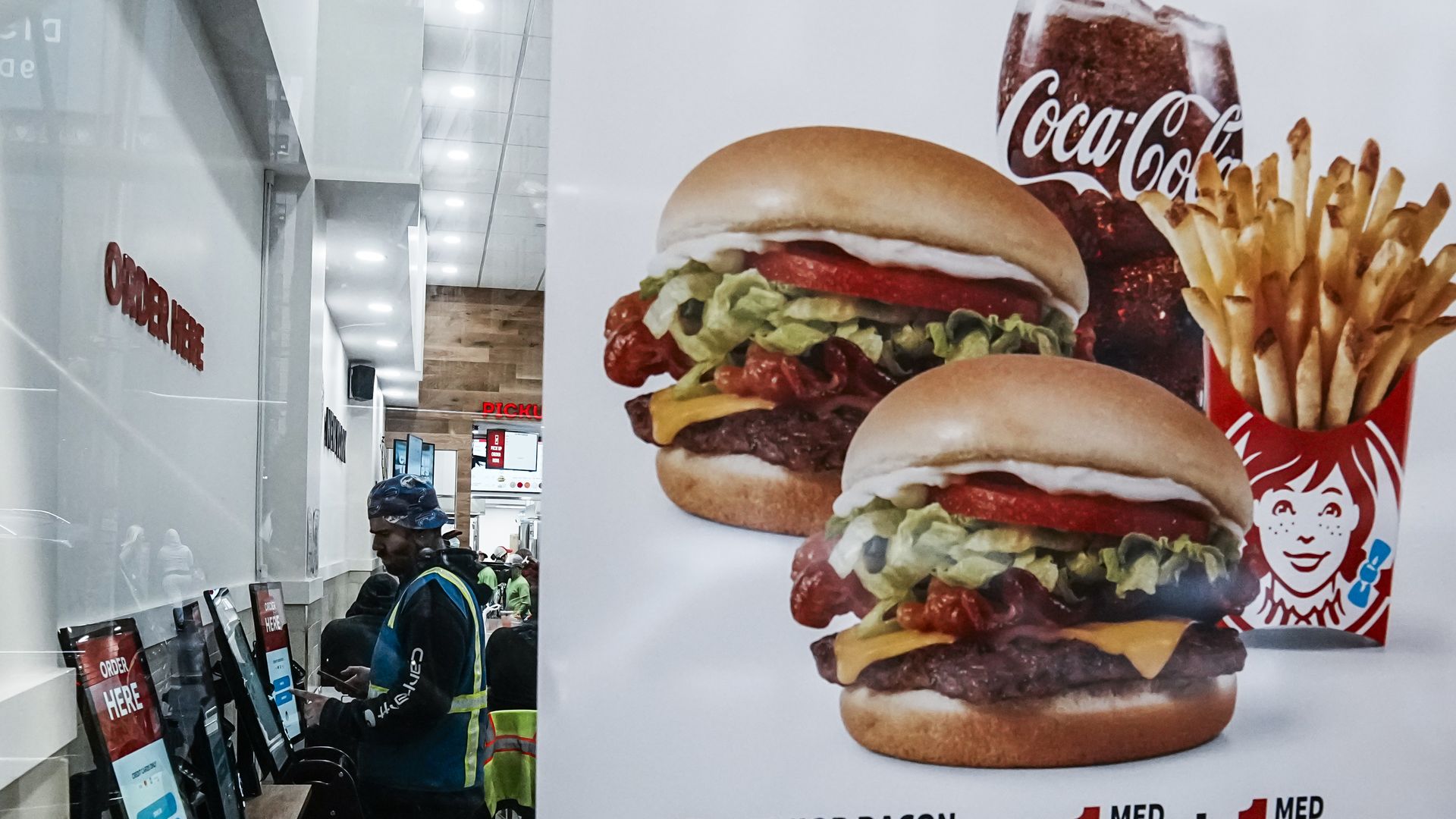 Fast food restaurant interior with a person using touchscreen order kiosks. Large advertisement shows two Junior Bacon Cheeseburgers with lettuce, tomato, cheese, plus medium fries and a Coca-Cola soft drink.