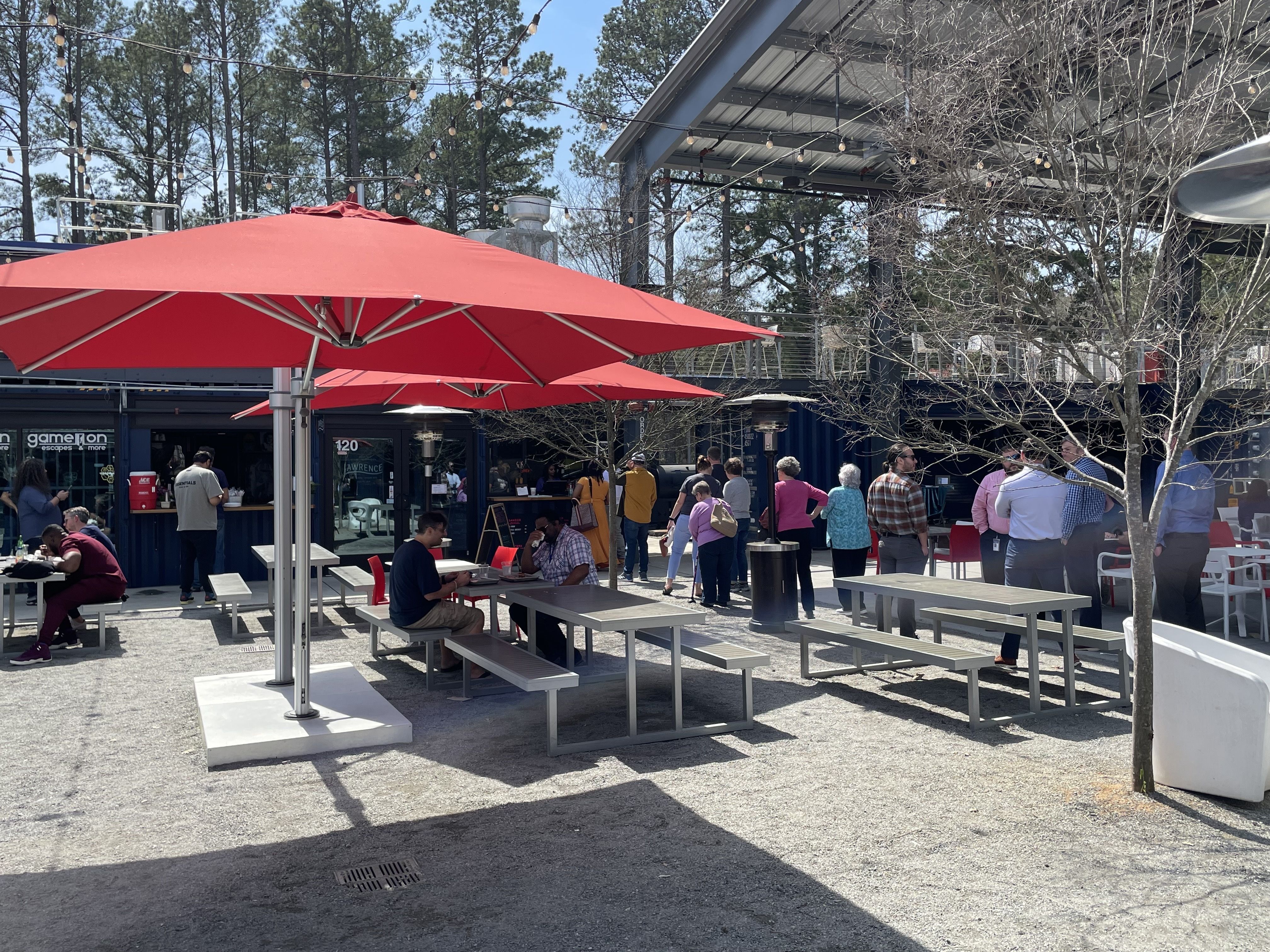 Outdoor seating area with gray tables and benches under large red umbrellas, people sitting and standing near blue container-style buildings, string lights overhead on a sunny day.