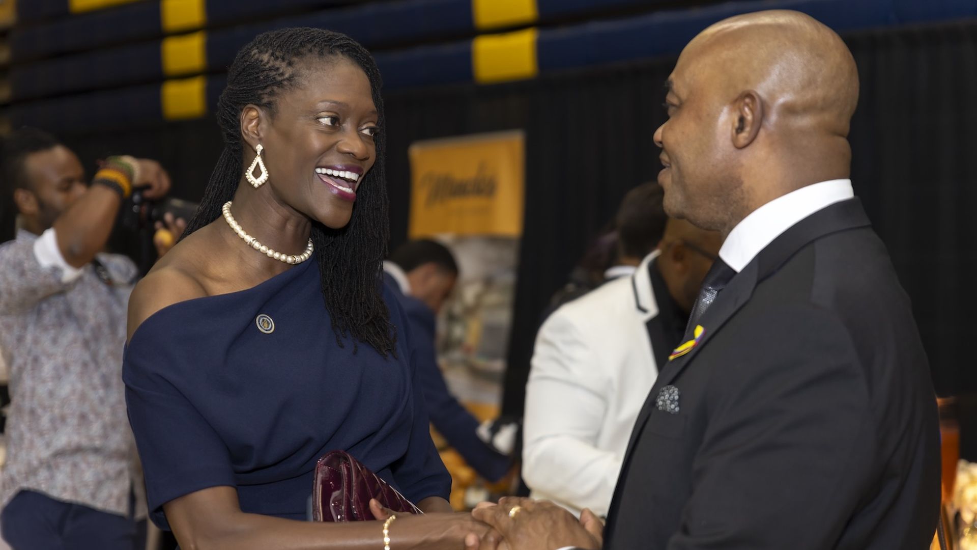 A woman in a navy blue, off-shoulder dress with a pearl necklace and earrings is smiling and shaking hands with a man in a formal suit and tie. She holds a maroon clutch. In the background, a person with a camera is taking photos, and blurred individuals are visible, including one in a white jacket.