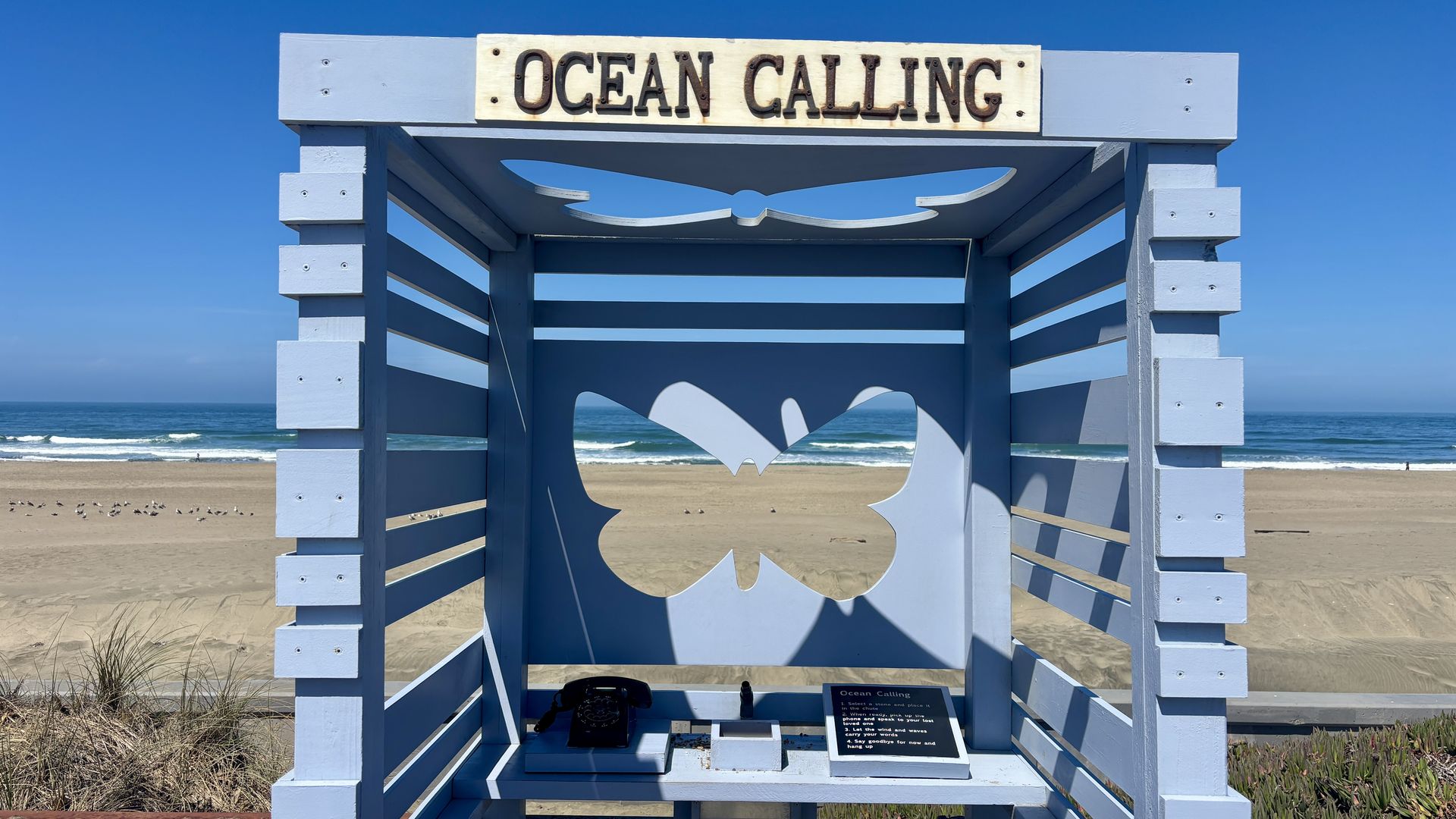 Light-blue wooden beach structure with a sign reading "Ocean Calling" at the top, a decorative cutout of a seagull inside, and a view of the sandy shore, ocean, and clear blue sky.