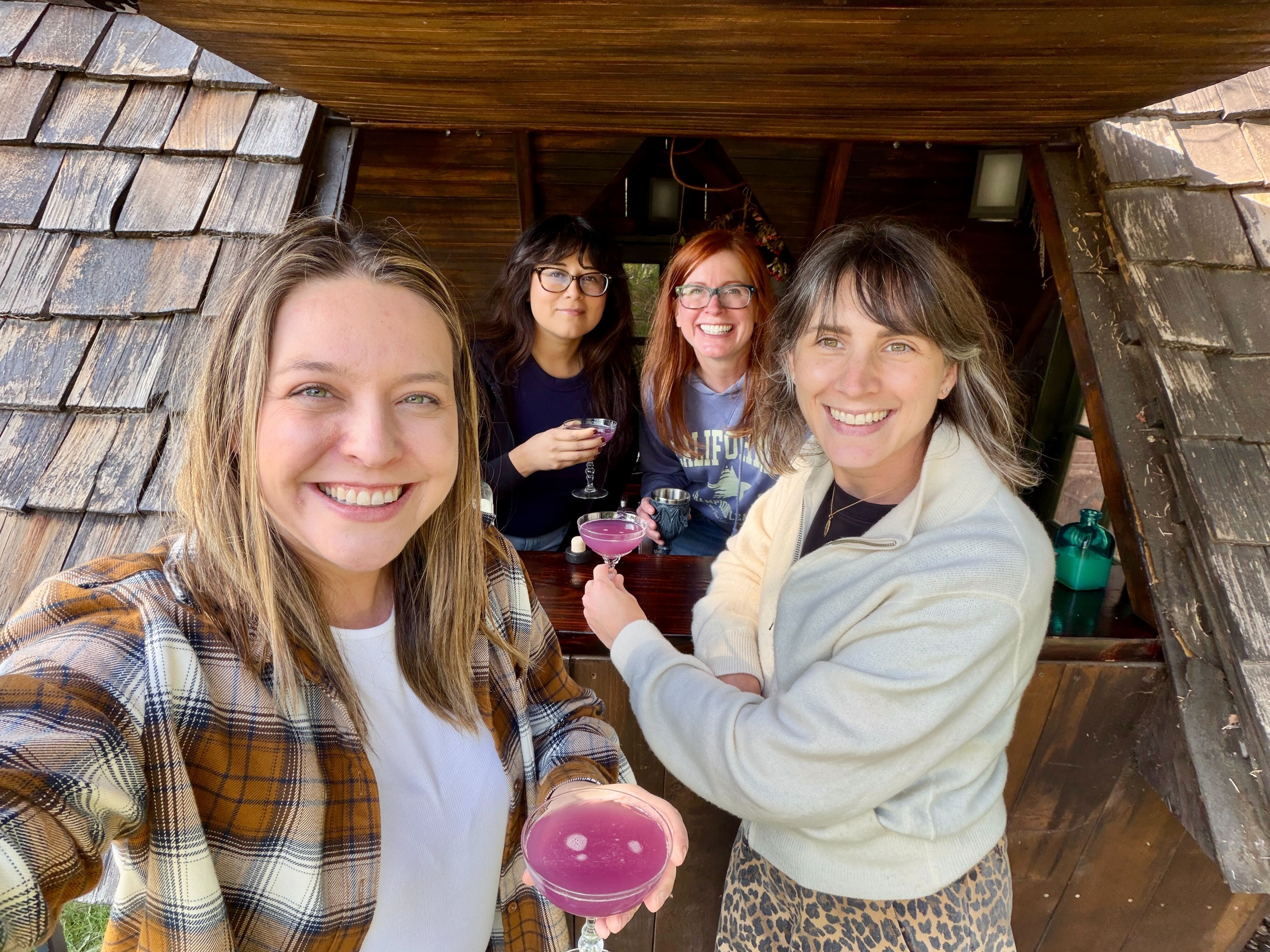 Four women hold cocktails at a wooden hut.