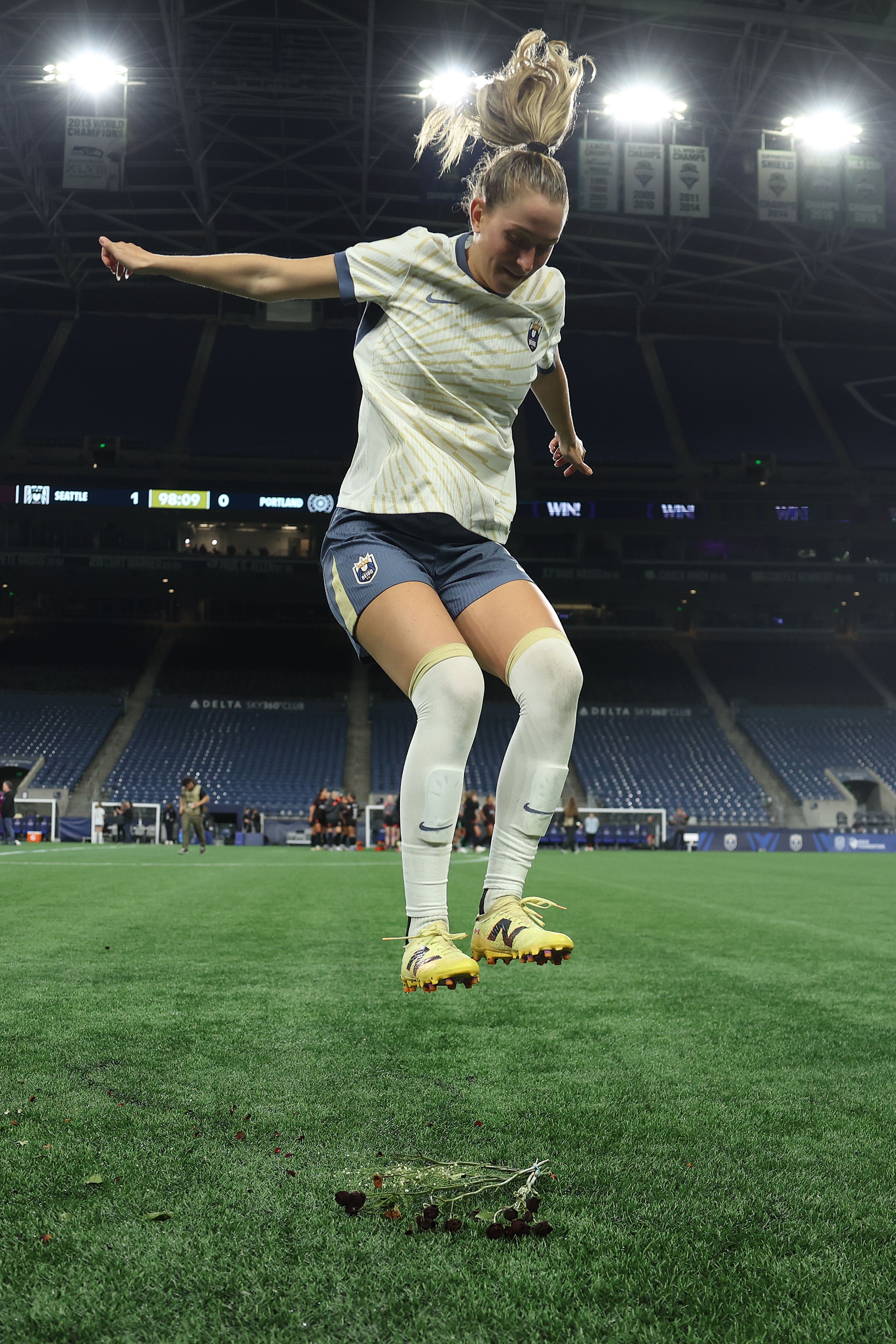 Female soccer player in white and navy uniform jumps above wilted flowers on green field under stadium lights with scoreboard showing Seattle 1, Portland 0, 98:09 minutes.