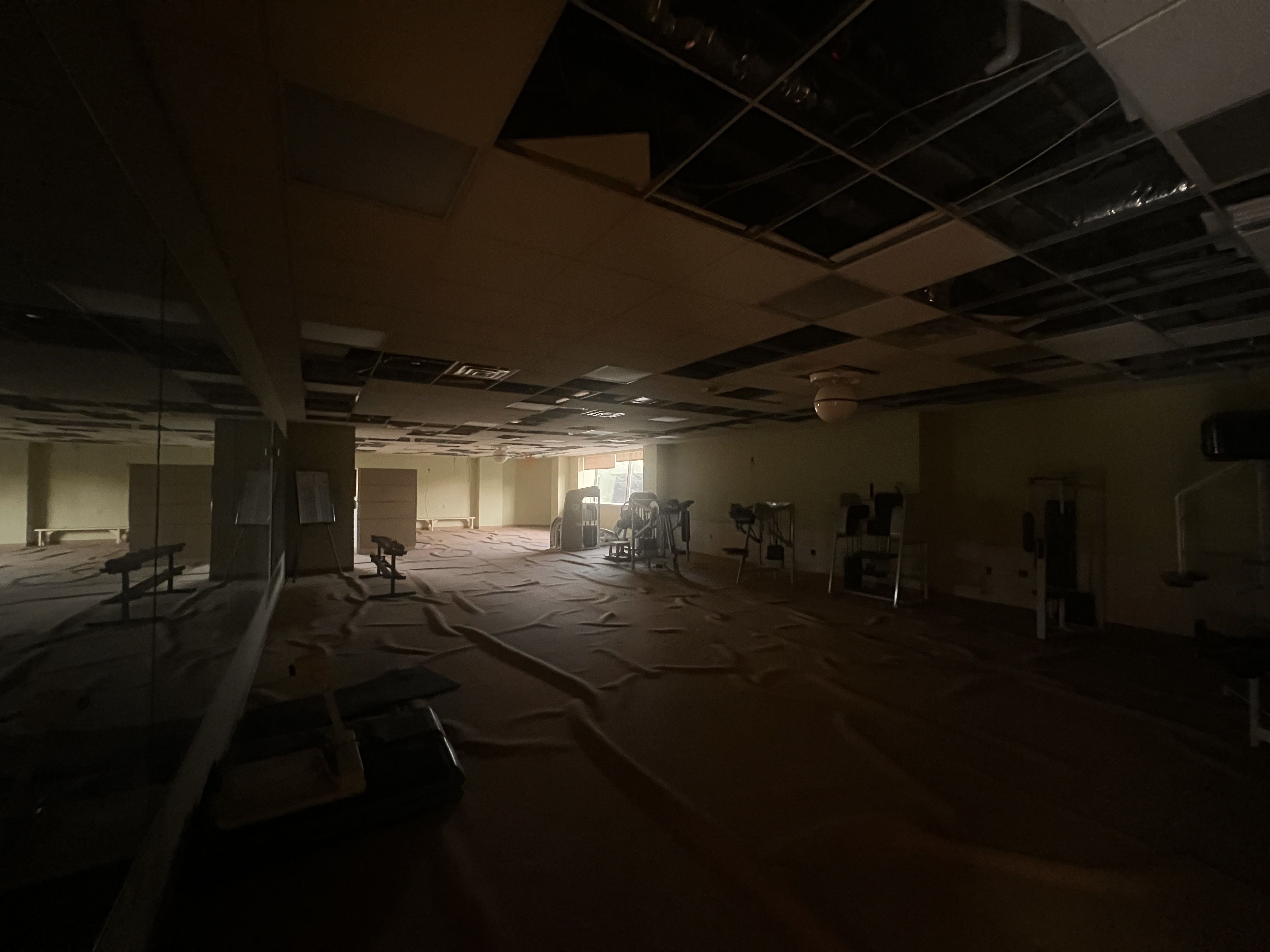 Dimly lit, abandoned gym with a damaged drop ceiling; many tiles missing, exposing dark ducts and wiring. Floor covered with protective sheets, with scattered benches and workout equipment.