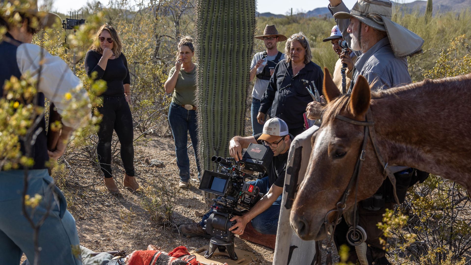 Film crew records scene in a desert with people standing around a man lying on the ground covered with a red blanket, a cameraman filming, and a brown horse in the foreground.